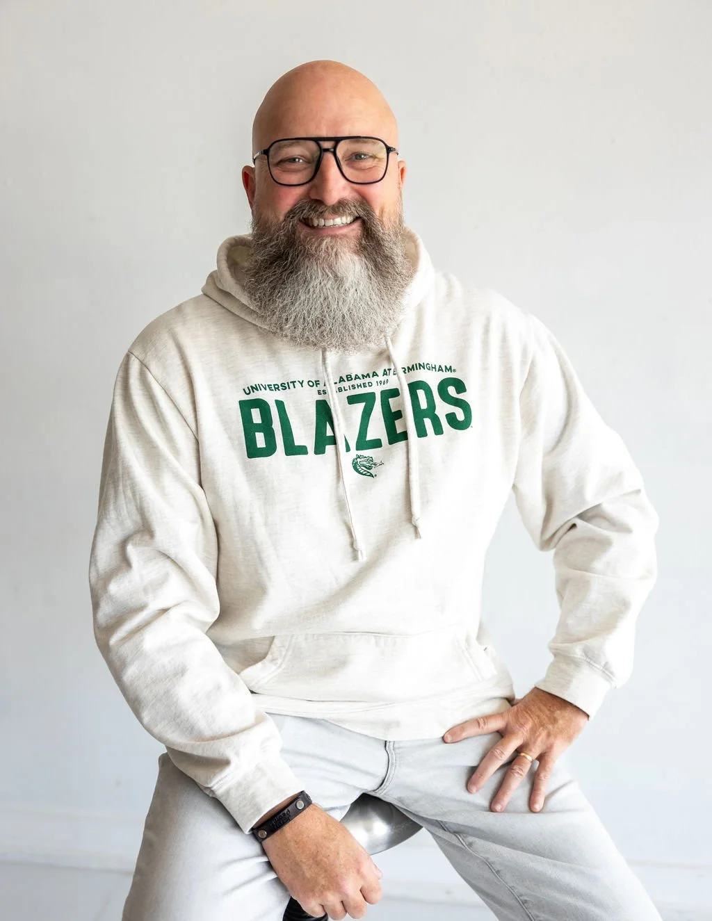 A smiling man with a bald head and a gray beard wearing glasses and a cream-colored hoodie with green text that says 'University of Alabama at Birmingham Blazers'. He is sitting on a stool, with one hand on his knee and the other resting on his hip, against a plain light background.