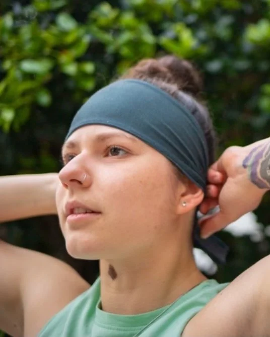Close-up of a woman with a headband, nose piercing, and tattoo, outdoors with greenery in the background, resting her arms behind her head.