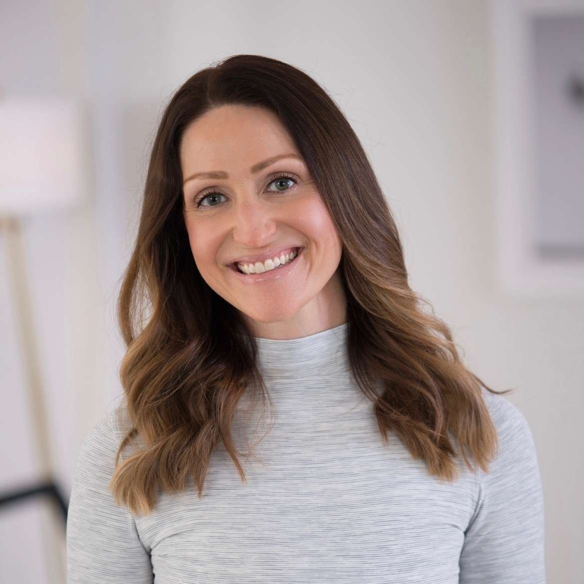 Smiling woman with long brown hair and blue eyes, wearing a light gray striped shirt, in a bright indoor setting.