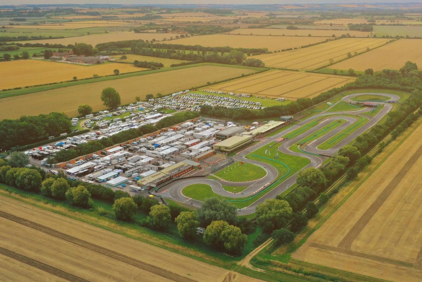 An aerial view of PFI go-kart racing track with multiple turns and green grassy areas, adjacent to a parking lot with numerous parked vehicles, surrounded by farmland and fields.