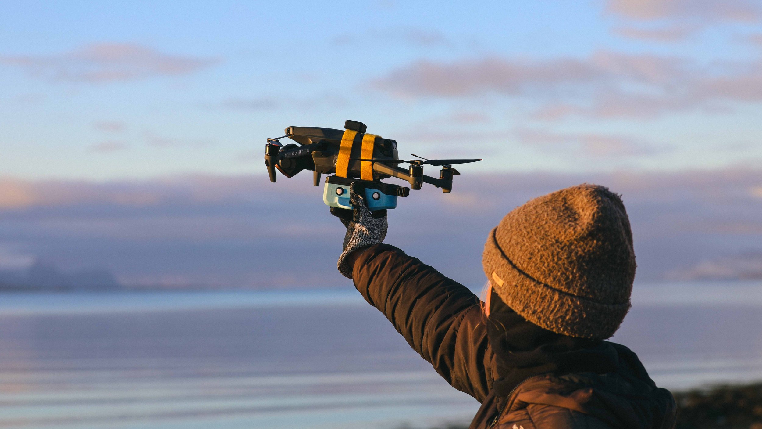 A person wearing a brown beanie and black jacket holding a drone in the air against a sky with clouds.