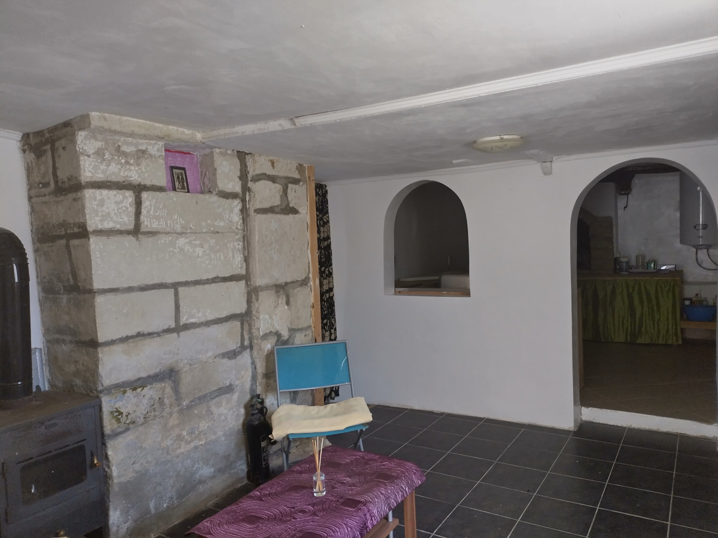 Interior room with stone stove, black pipe, chair, table, and arched wall openings; tiled floor and white ceiling.