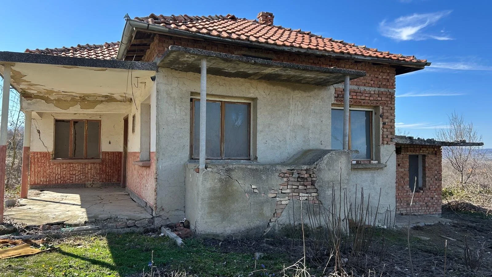 Abandoned house with cracked walls and broken windows, surrounded by overgrown vegetation, against a clear blue sky.