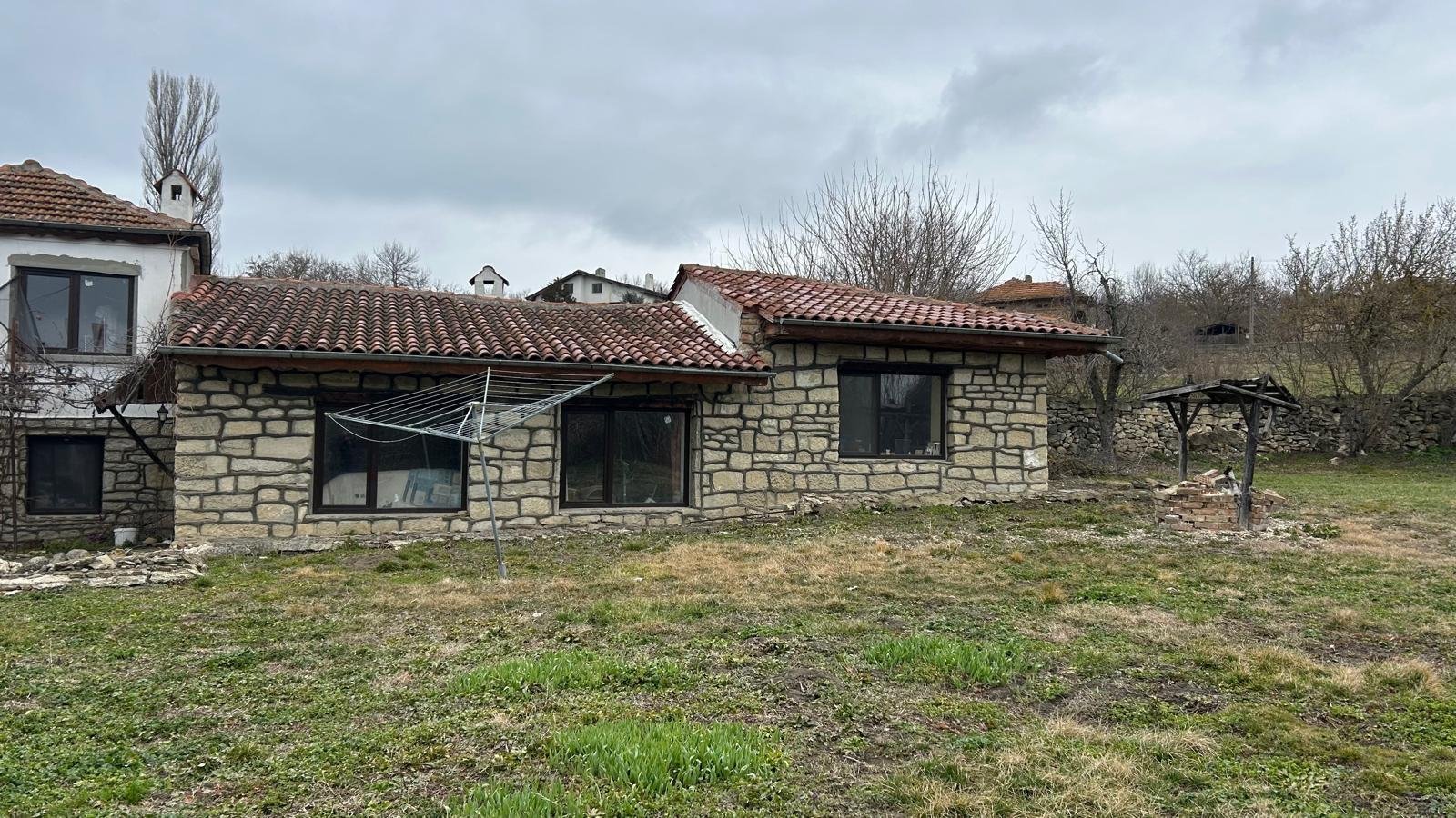 A stone house with red tiled roof in a rural area with grass and a well