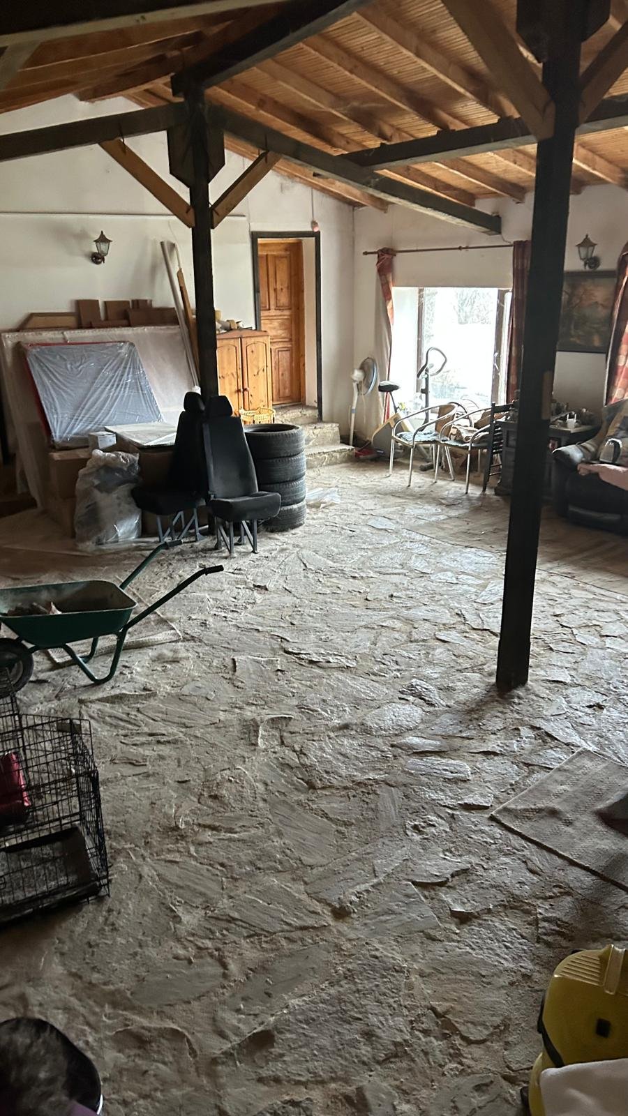 Interior of a rustic, cluttered room with exposed wooden beams, stone floor, a stack of tires, chairs, a wheelbarrow, a cupboard, and various household items.
