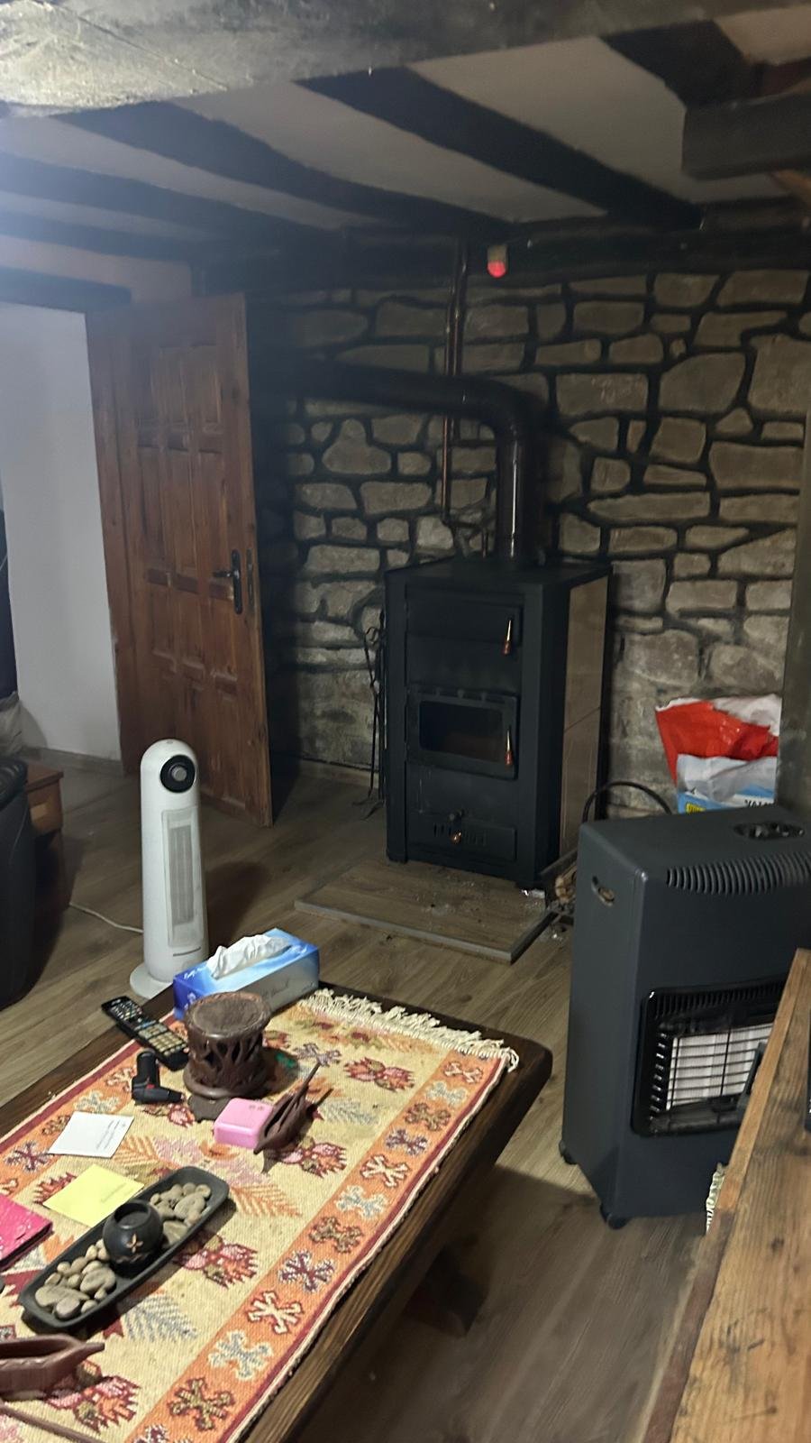 Cozy living room with a rustic wood-burning stove against a stone wall, a wooden coffee table with colorful tablecloth, tissue box, decorative items, and a tower fan.