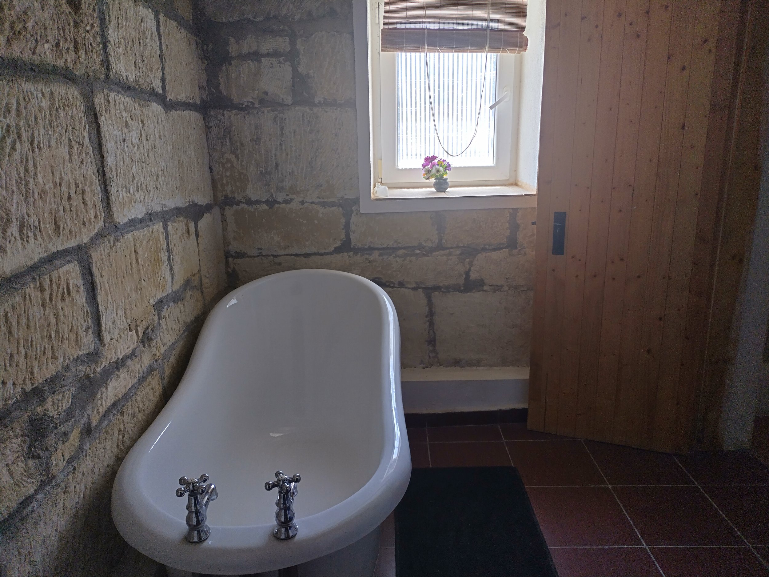 Bathroom with a white bathtub, stone walls, a wooden door, and a small window with a blind and a vase of flowers on the sill.