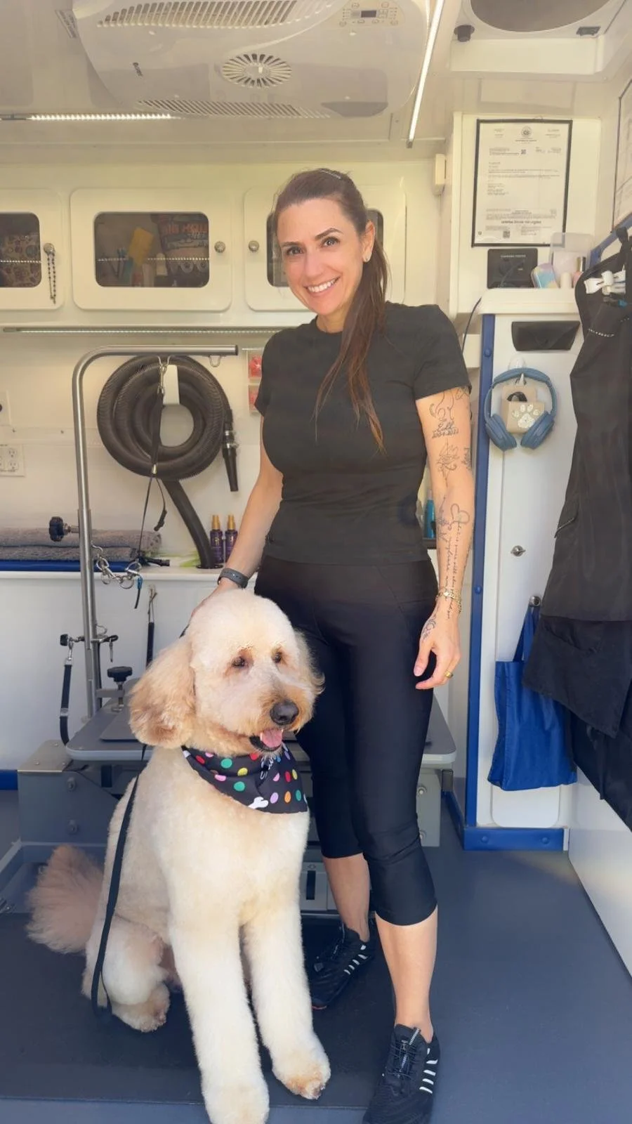 A woman with tattoos and dark hair smiling while holding a leash attached to a large, fluffy, light-colored dog with a colorful bandana around its neck, inside what appears to be a mobile grooming van.
