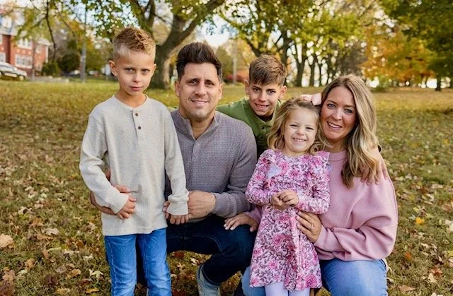 Family of five outdoor in a park with autumn trees, smiling for a photo.