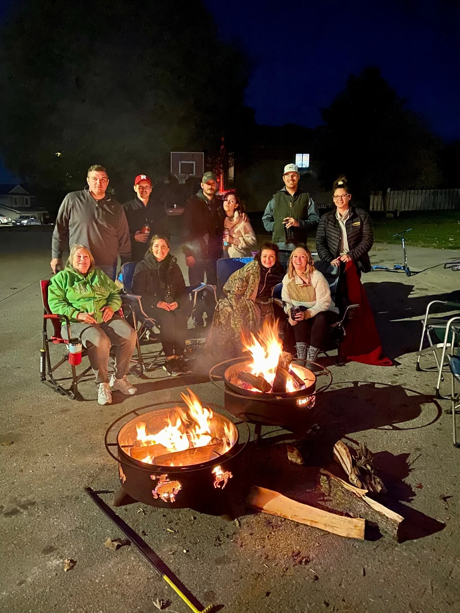 Group of people gathered around a campfire outdoors at night, with some sitting in chairs and some standing, with a basketball hoop and houses in the background.