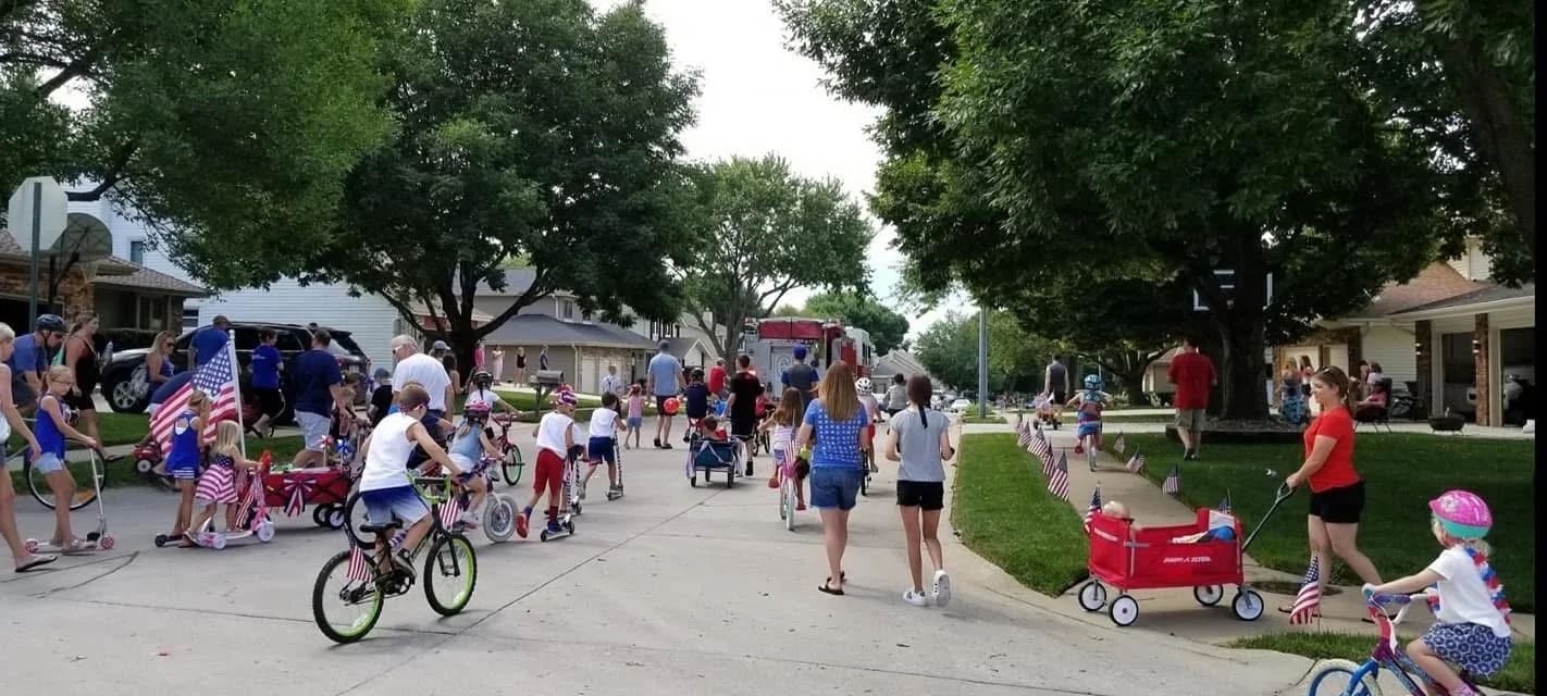 A neighborhood parade with children riding bikes, playing, and walking, decorated with American flags, adults present, trees and houses in the background, and a fire truck in the distance.