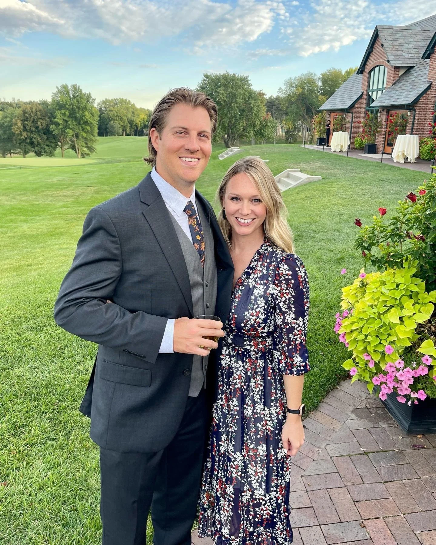 A man in a dark suit and a woman in a floral dress smiling outdoors on a manicured lawn with a brick building and a tree-lined background.