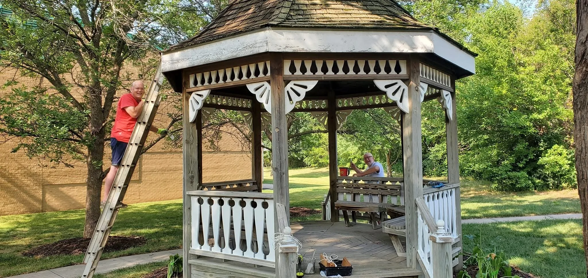 Two men, one climbing a ladder on the left and another sitting at a picnic table inside a wooden gazebo, in a park with green grass and trees.
