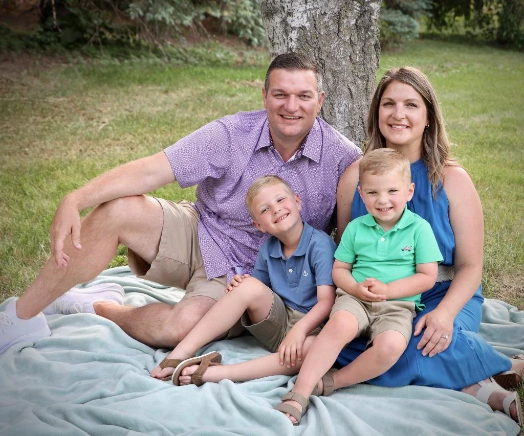 A family of four sitting on a blanket outdoors under a tree, smiling at the camera. The father, with short dark hair, is wearing a purple shirt and khaki shorts. The mother, with shoulder-length light brown hair, is wearing a blue sleeveless dress. Their two young boys, with blond hair, are sitting in front; one in a blue shirt and shorts, the other in a green polo shirt and khaki shorts.