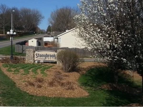 A landscaped area with budding bushes and flowering trees, a brick sign that reads 'Stonybrook', and residential houses in the background under a clear blue sky.