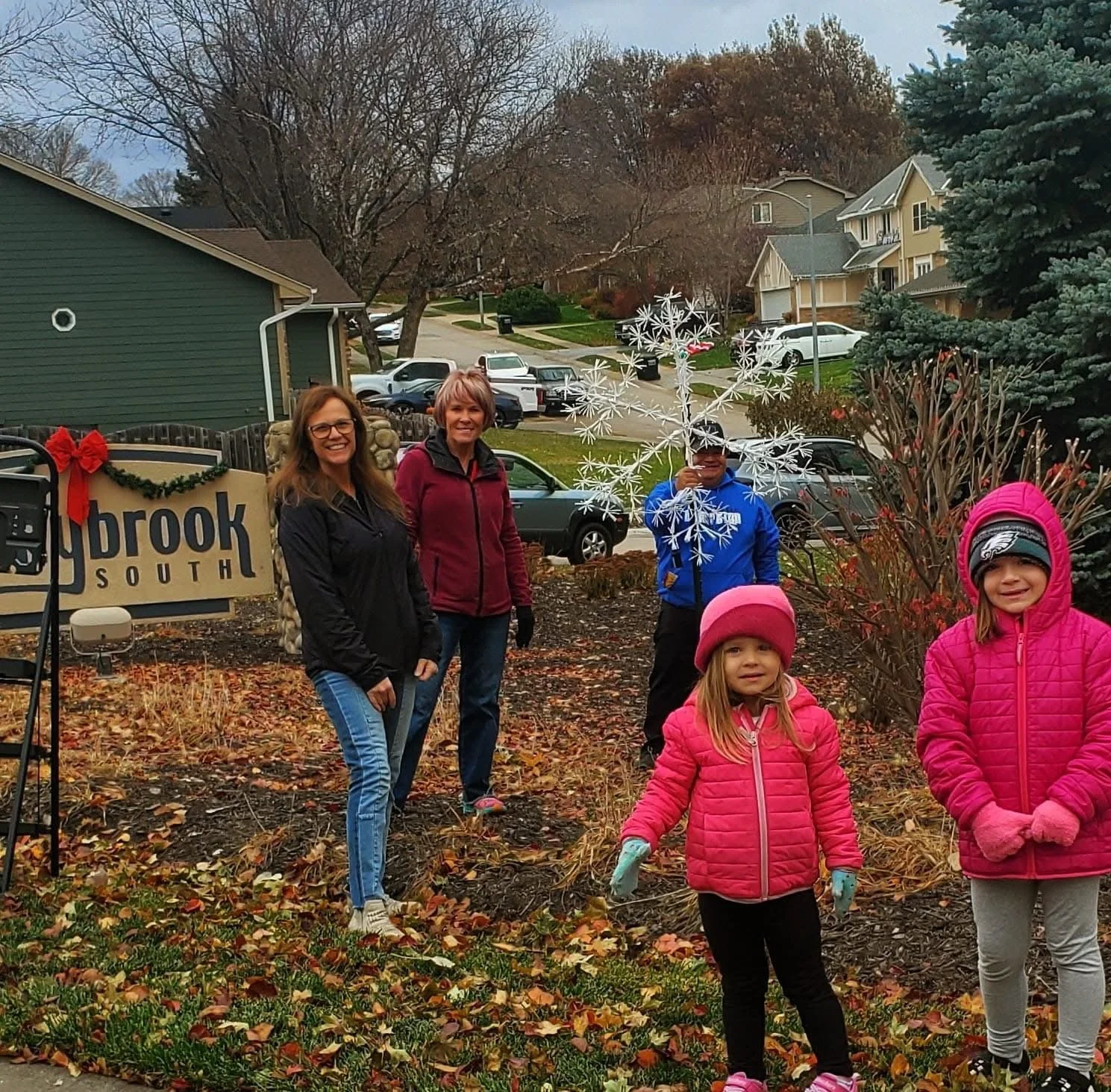 Group of people, including two adults and three children, planting or tending to a garden in a neighborhood yard during fall, with leafless trees, parked cars, and houses in the background.