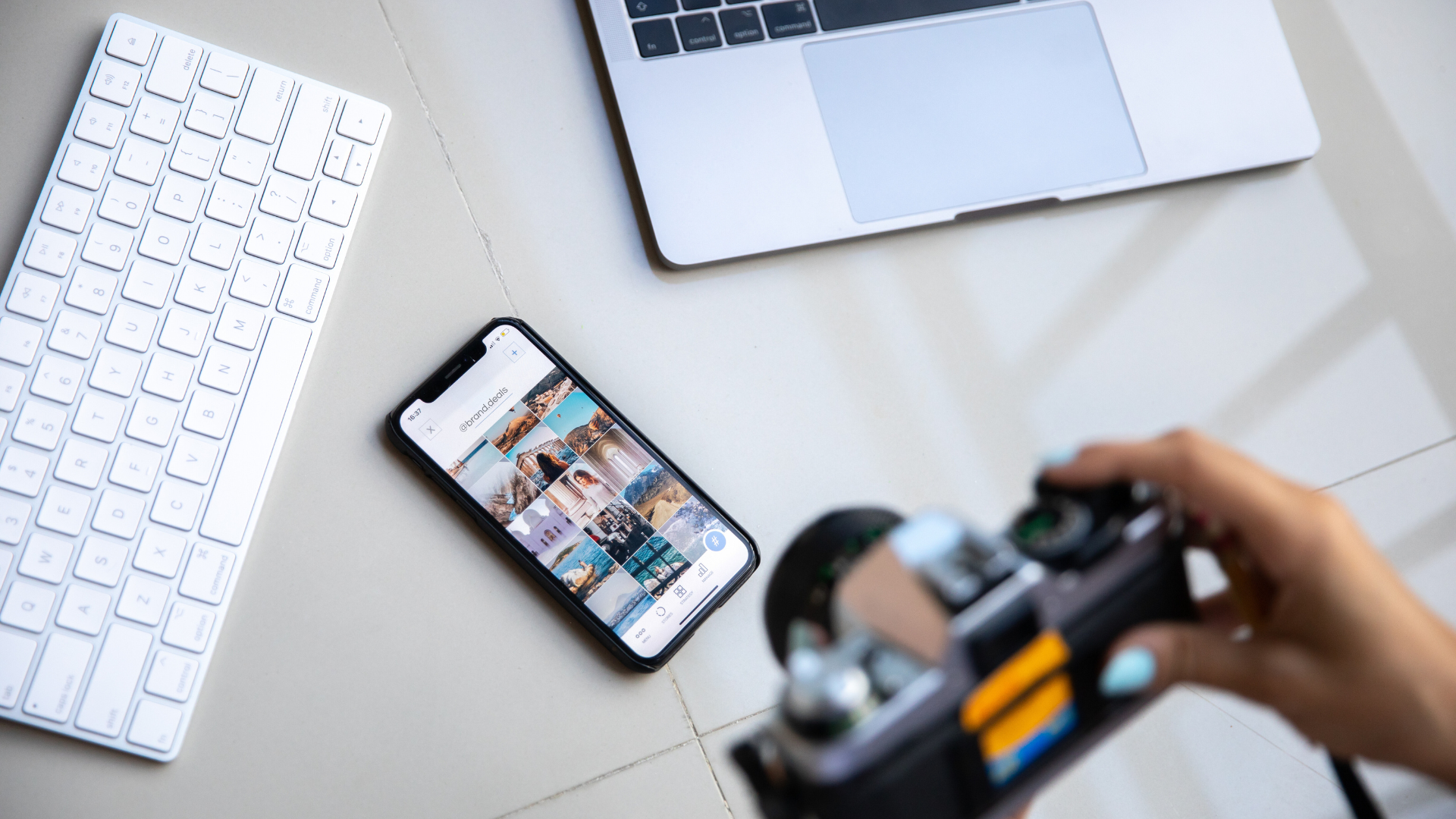 A modern desk setup featuring a white keyboard, a partially visible silver laptop, a smartphone displaying a grid of nature and travel photos, and a person holding a camera. The scene is on a light-colored tiled surface.