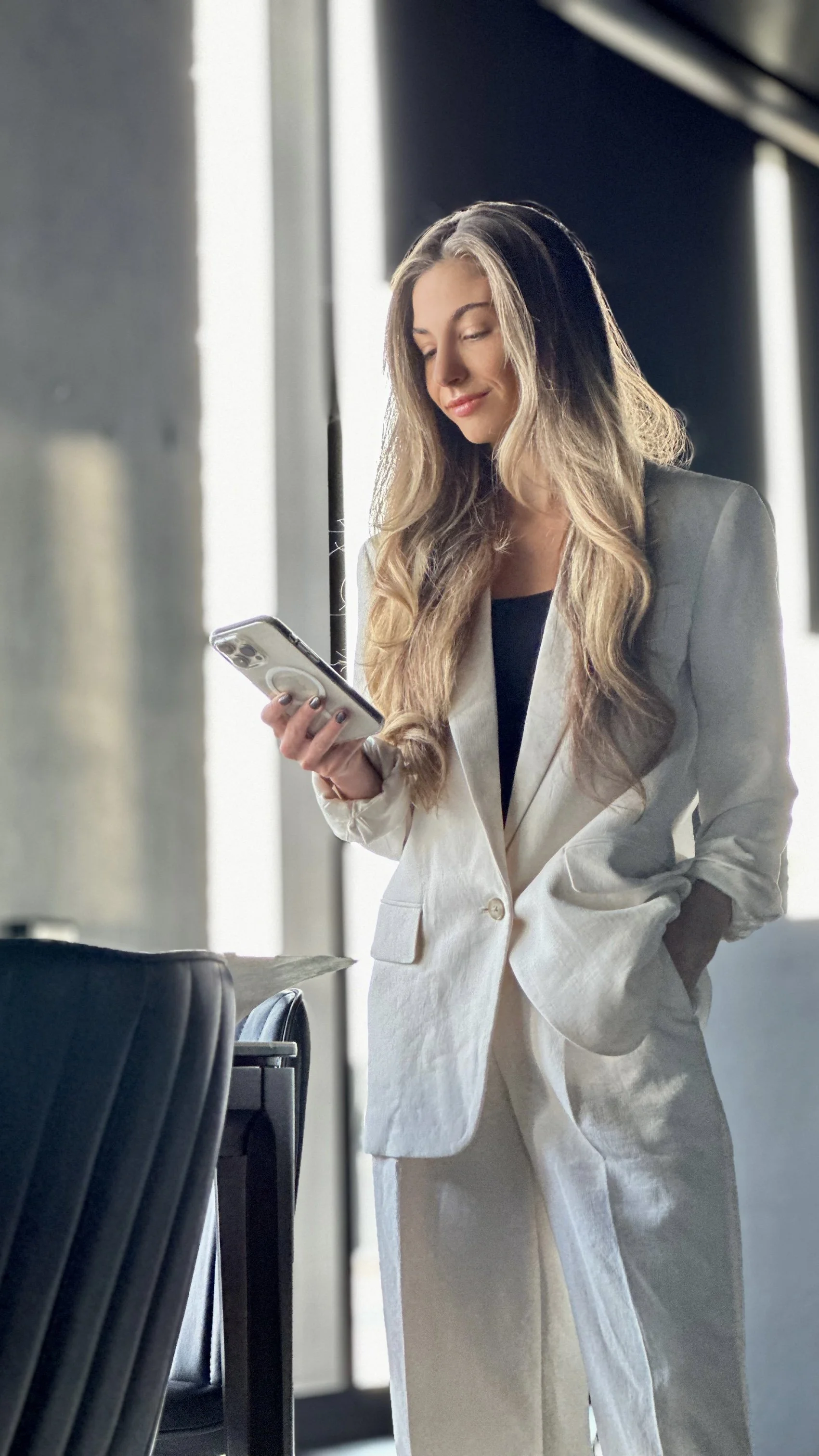 Marli, a lawyer with long hair in a white suit holding a smartphone, standing next to a chair in a modern indoor setting.