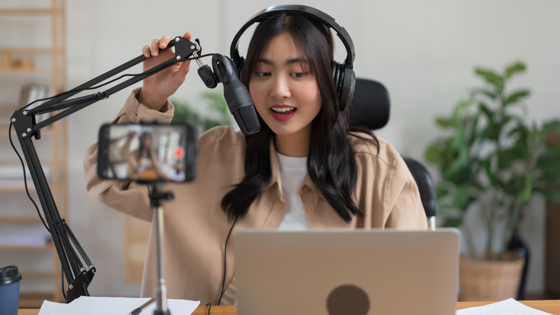 A young woman recording a podcast or video, wearing headphones, speaking into a microphone, with a smartphone on a tripod capturing her, sitting at a desk with papers and a laptop, in a bright room with plants in the background.