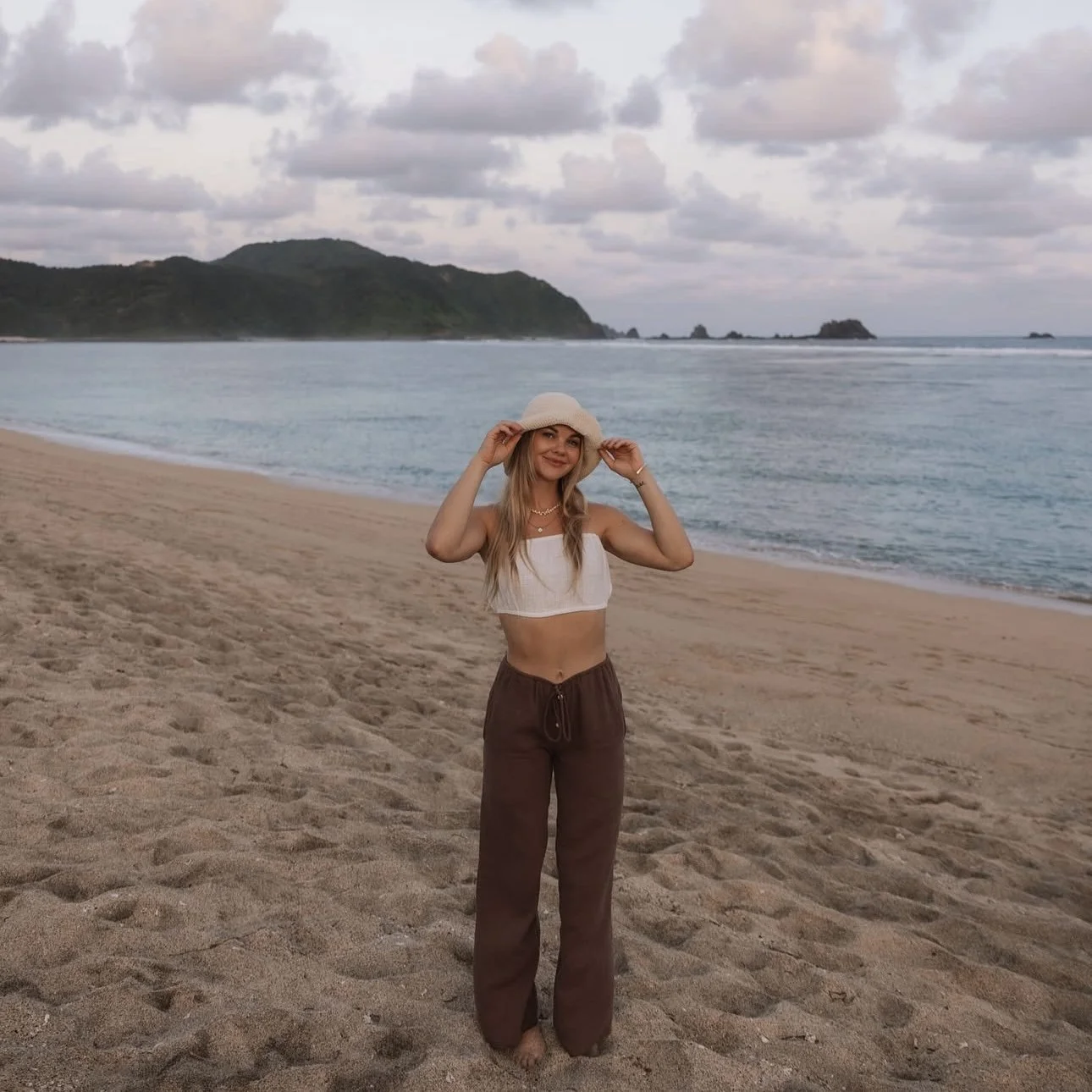 Woman standing on a sandy beach, holding a wide-brimmed hat, with ocean waves, rocky islands, and a mountain in the background.