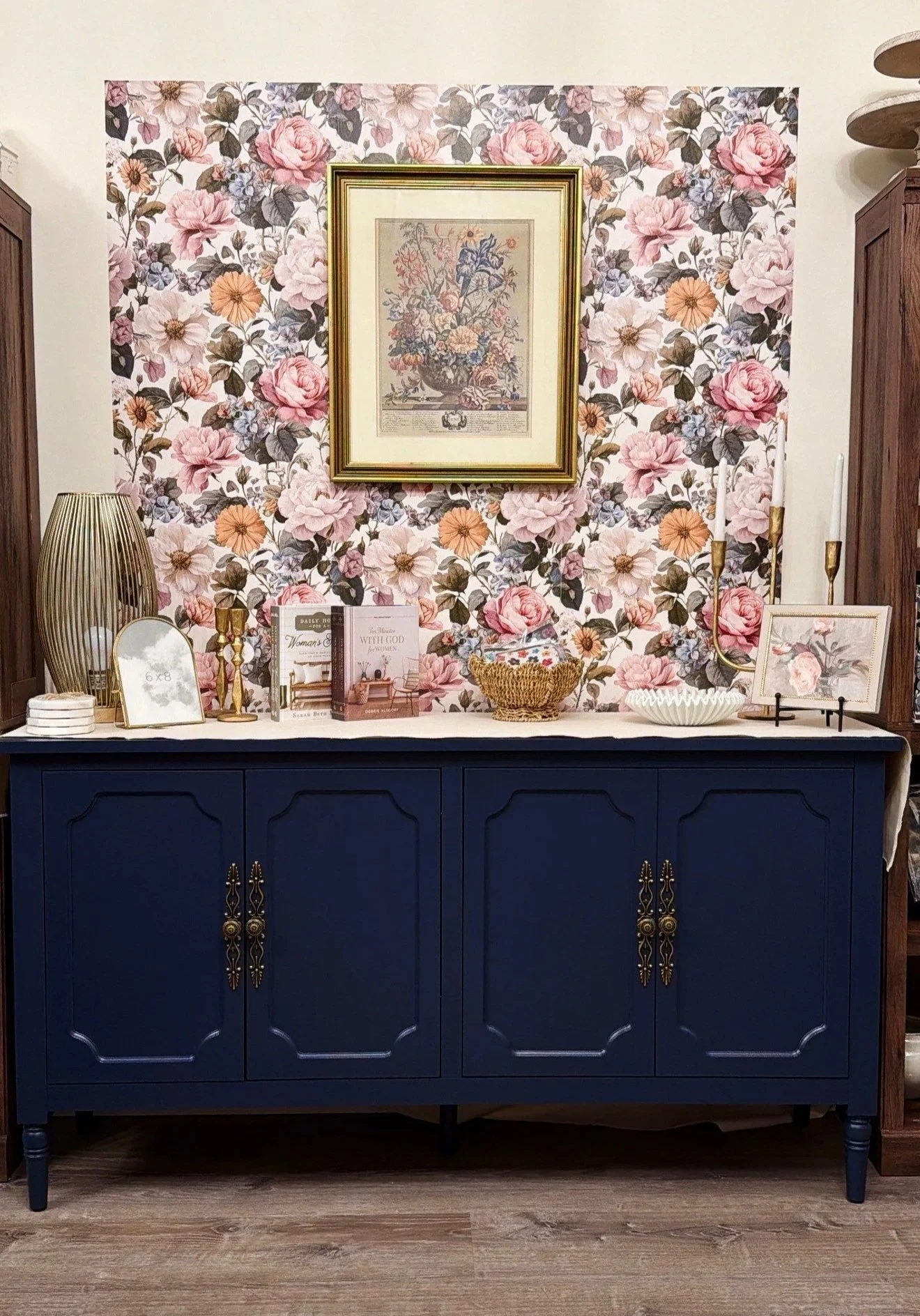 A navy blue sideboard decorated with books, vases, picture frames, and candlesticks, against a floral wallpaper background with pink and peach flowers.