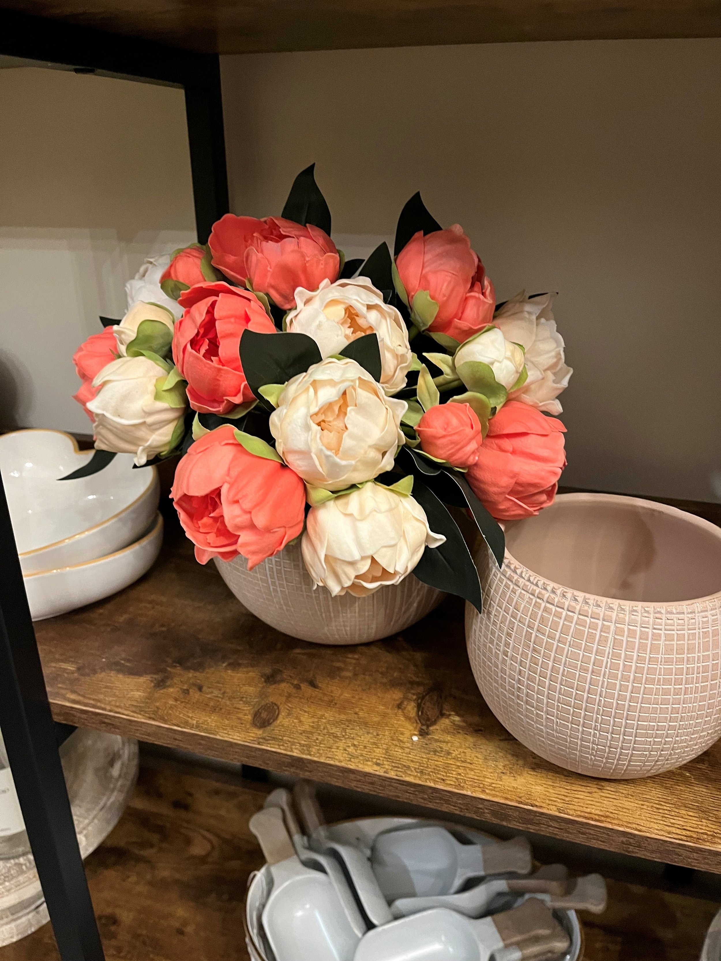 A bouquet of pink and white peonies arranged in a cream-colored textured ceramic vase, placed on a wooden shelf.