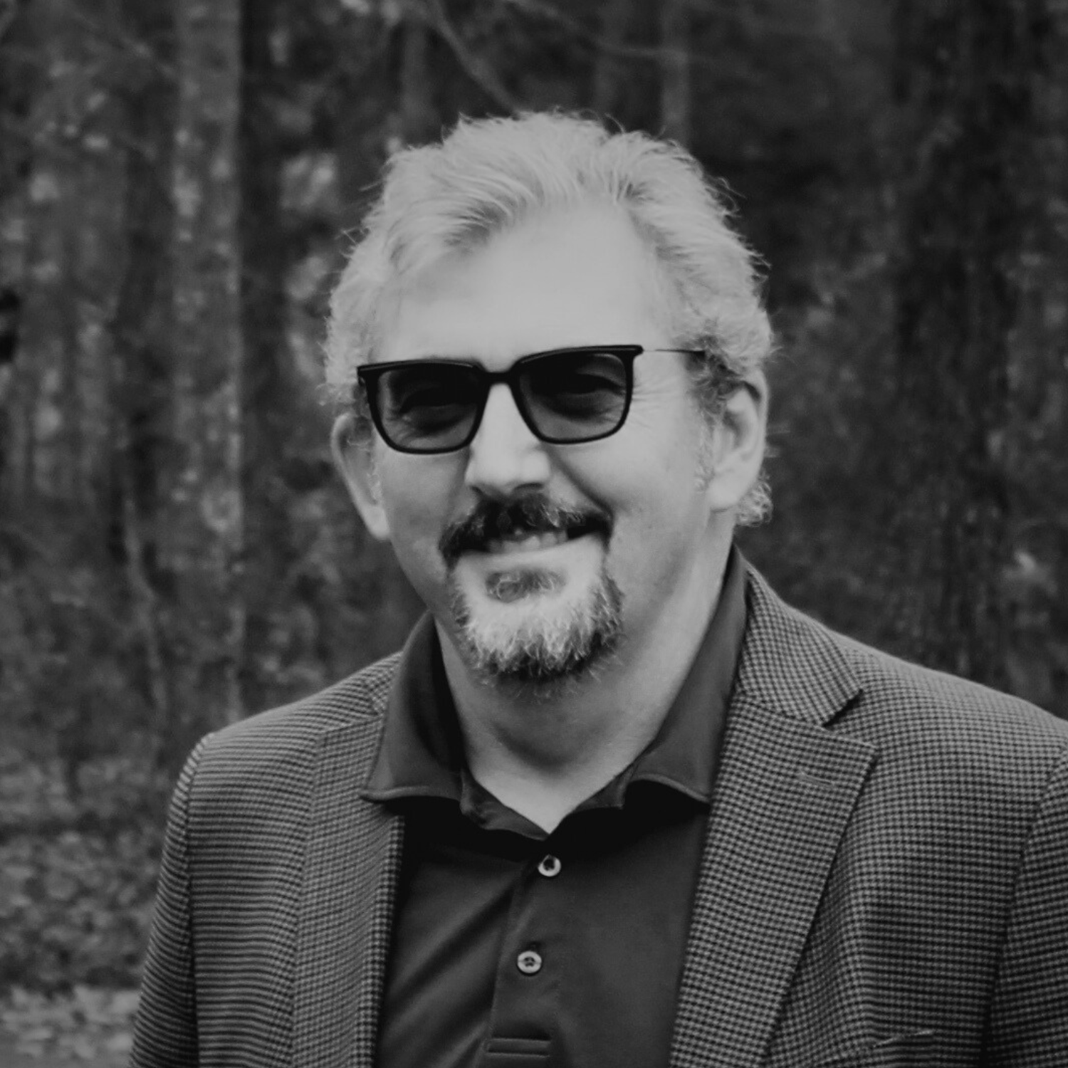 A middle-aged man with short, gray hair and a goatee stands outdoors in front of autumn-colored trees, wearing a beige blazer over a checked shirt.