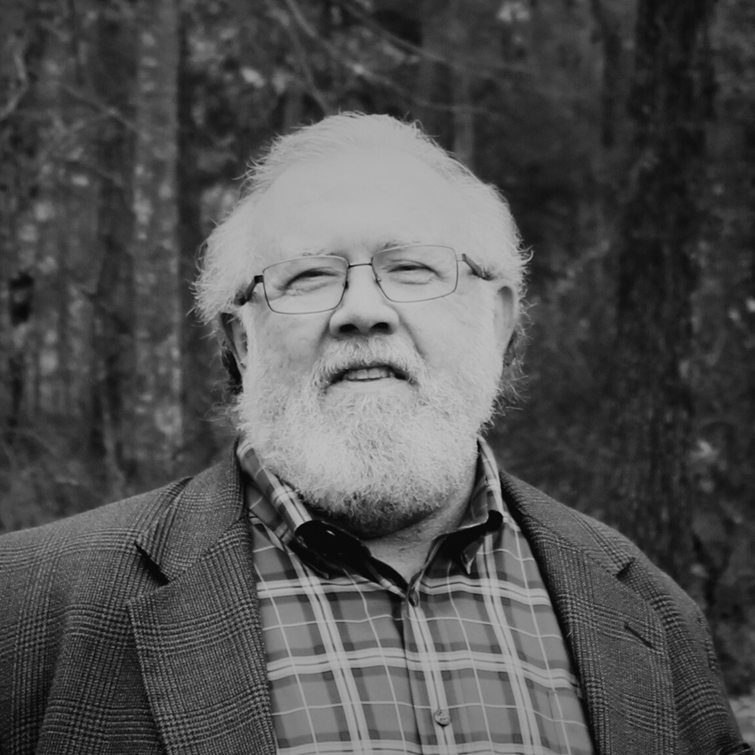 A man with glasses, a beard, and gray hair standing outdoors among trees, wearing a black blazer and a blue plaid shirt.