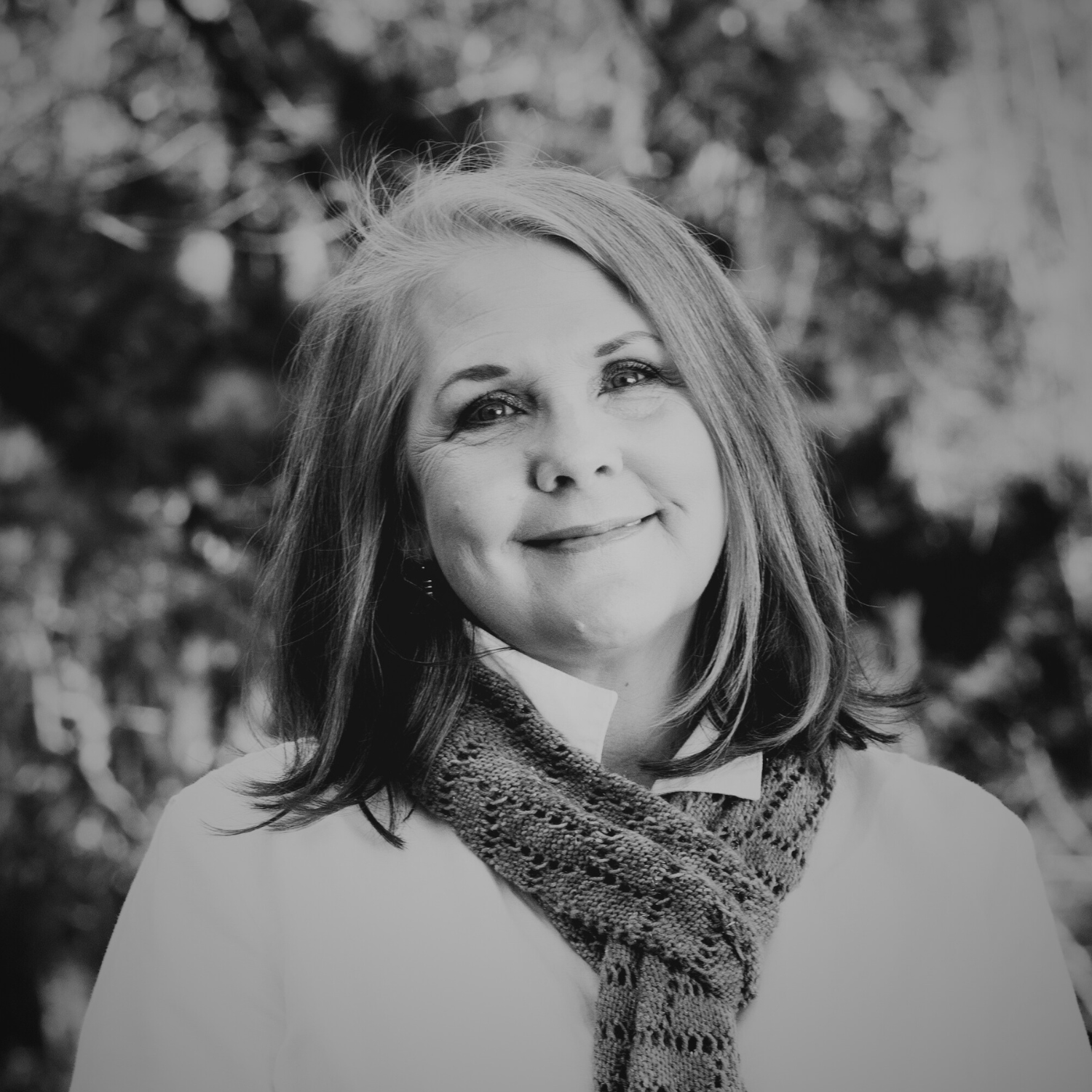 A woman with shoulder-length light brown hair, wearing a white button-up shirt, a green patterned scarf, and black pants, standing outdoors in a park during daytime with trees and autumn foliage in the background.