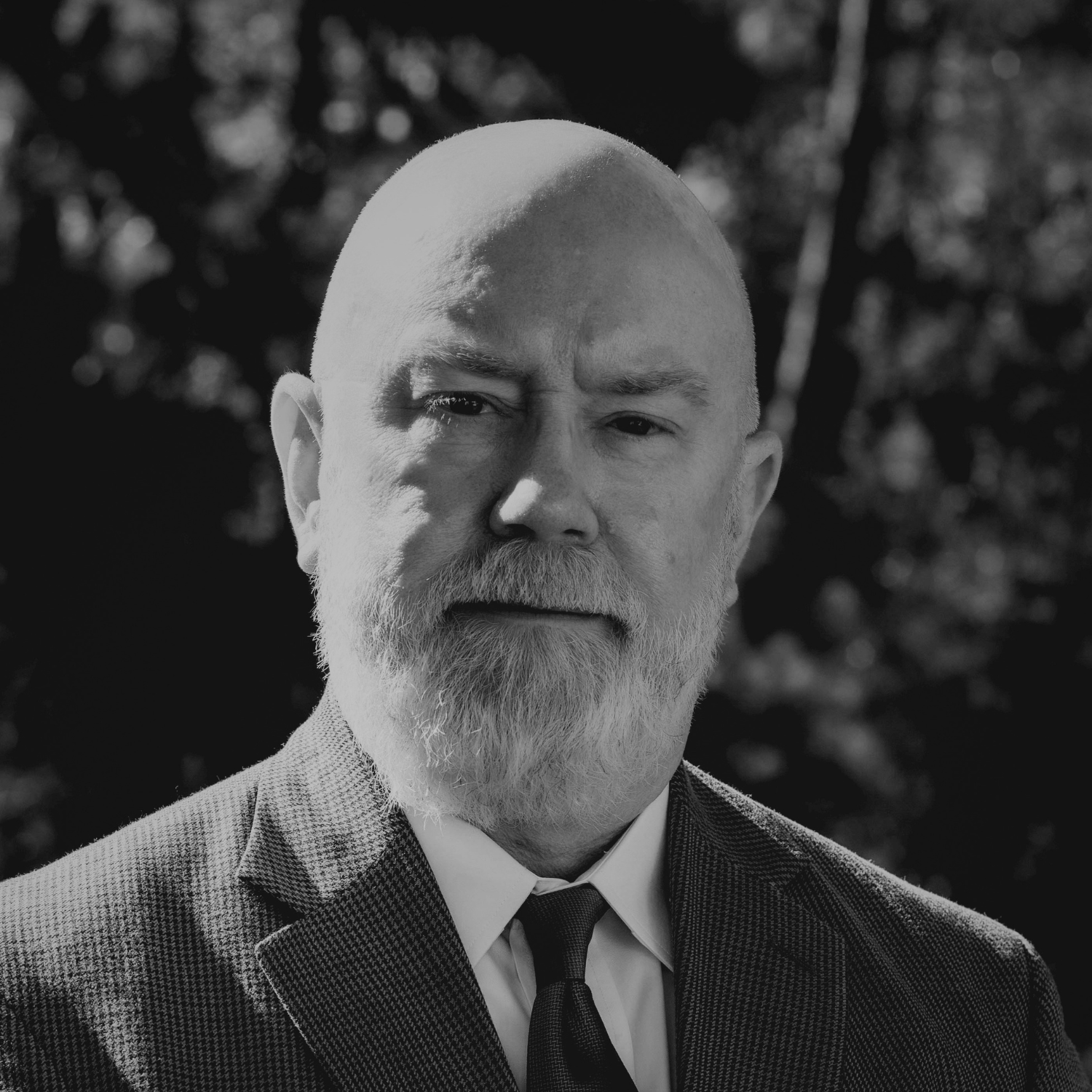 A man with a white beard and bald head stands outdoors in front of trees with autumn-colored leaves, wearing a brown plaid blazer, white shirt, and red tie.