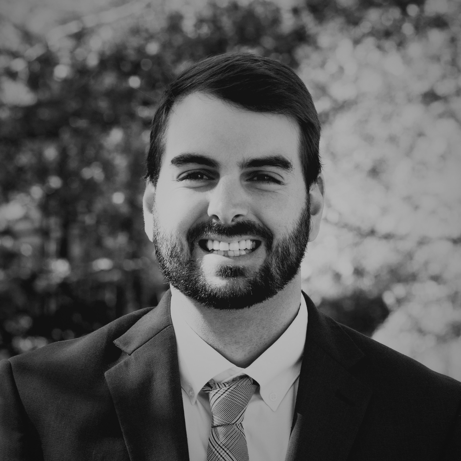 A smiling man in a navy suit, white dress shirt, and striped tie standing outdoors with fall foliage in the background.