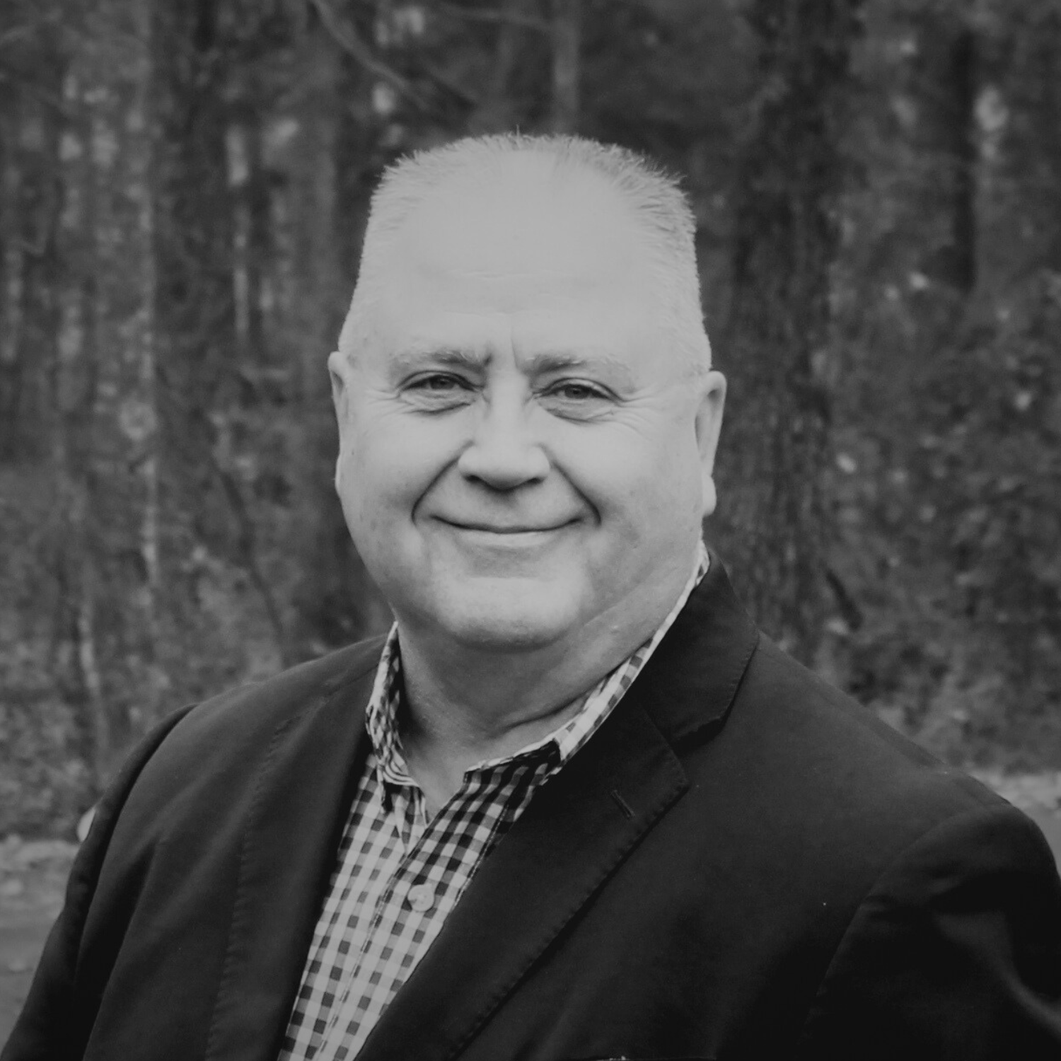 A middle-aged man in a tan suit with a white shirt and patterned tie standing outdoors in front of trees with autumn leaves.