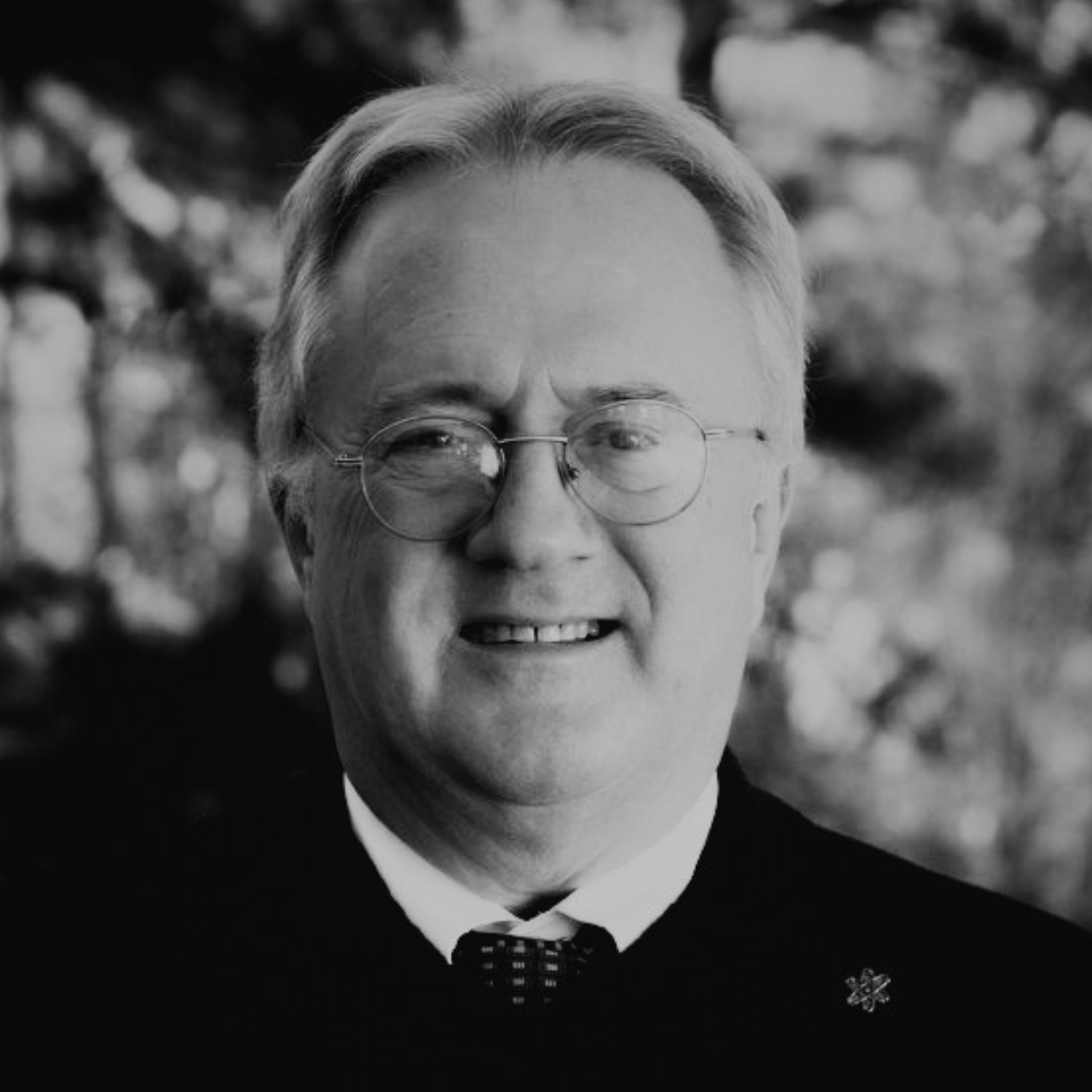 A man wearing glasses, a white shirt, black tie, and black jacket standing outdoors in front of trees.