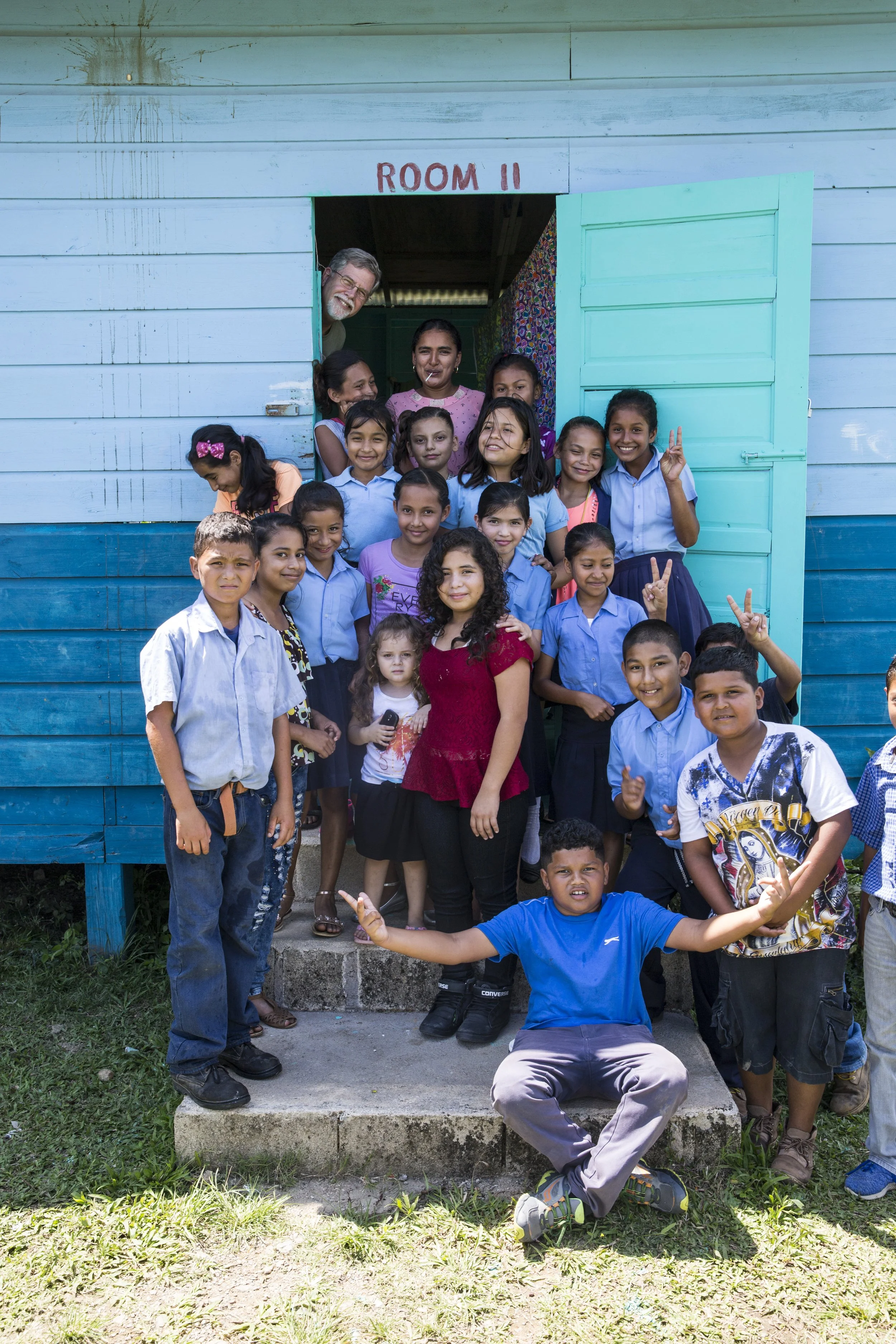 A group of children and two adults standing outside a blue wooden building labeled 'Room II'. The children are smiling, some making peace signs, and are dressed in casual clothes and school uniforms. One boy in a blue shirt is sitting in front with arms spread wide.