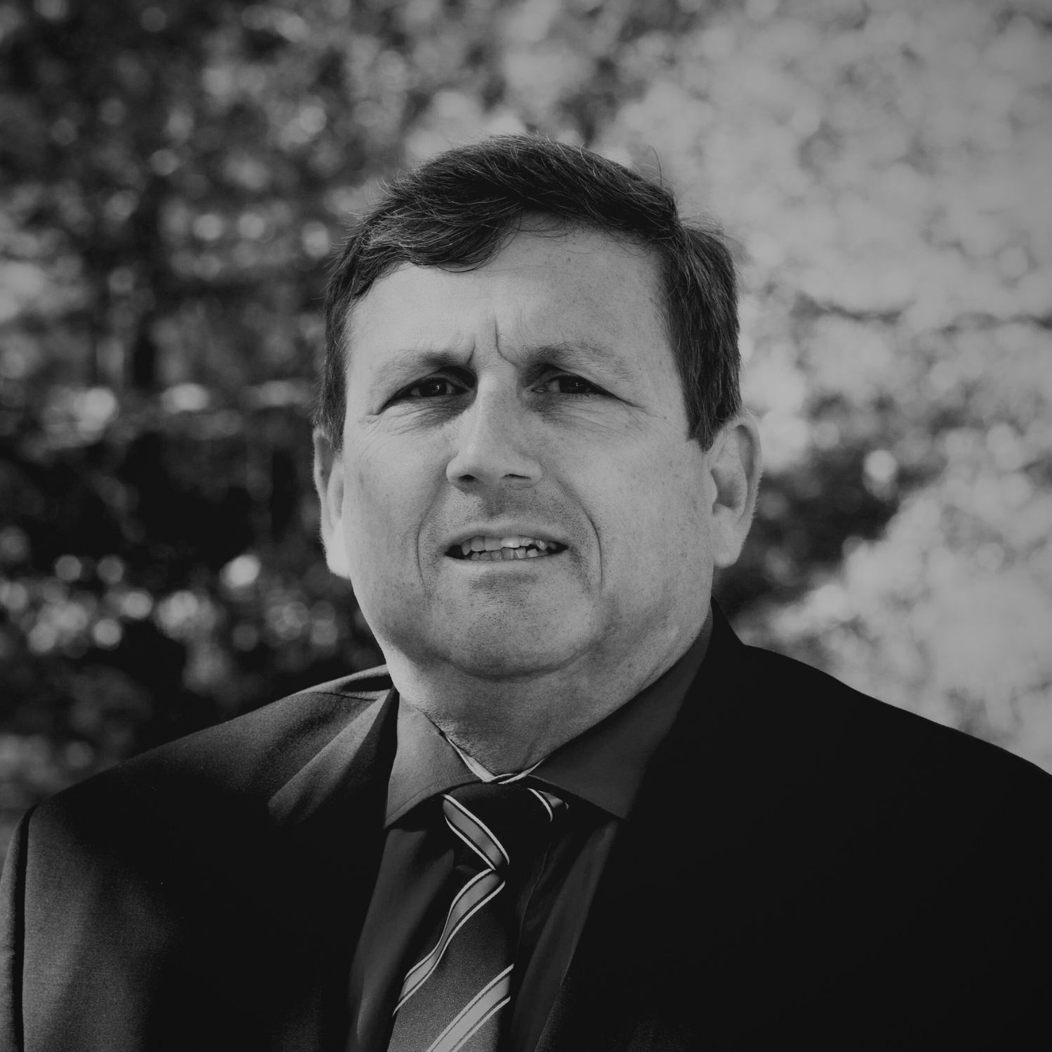 A middle-aged man in a black suit, blue shirt, and striped tie standing outdoors with autumn-colored trees in the background.
