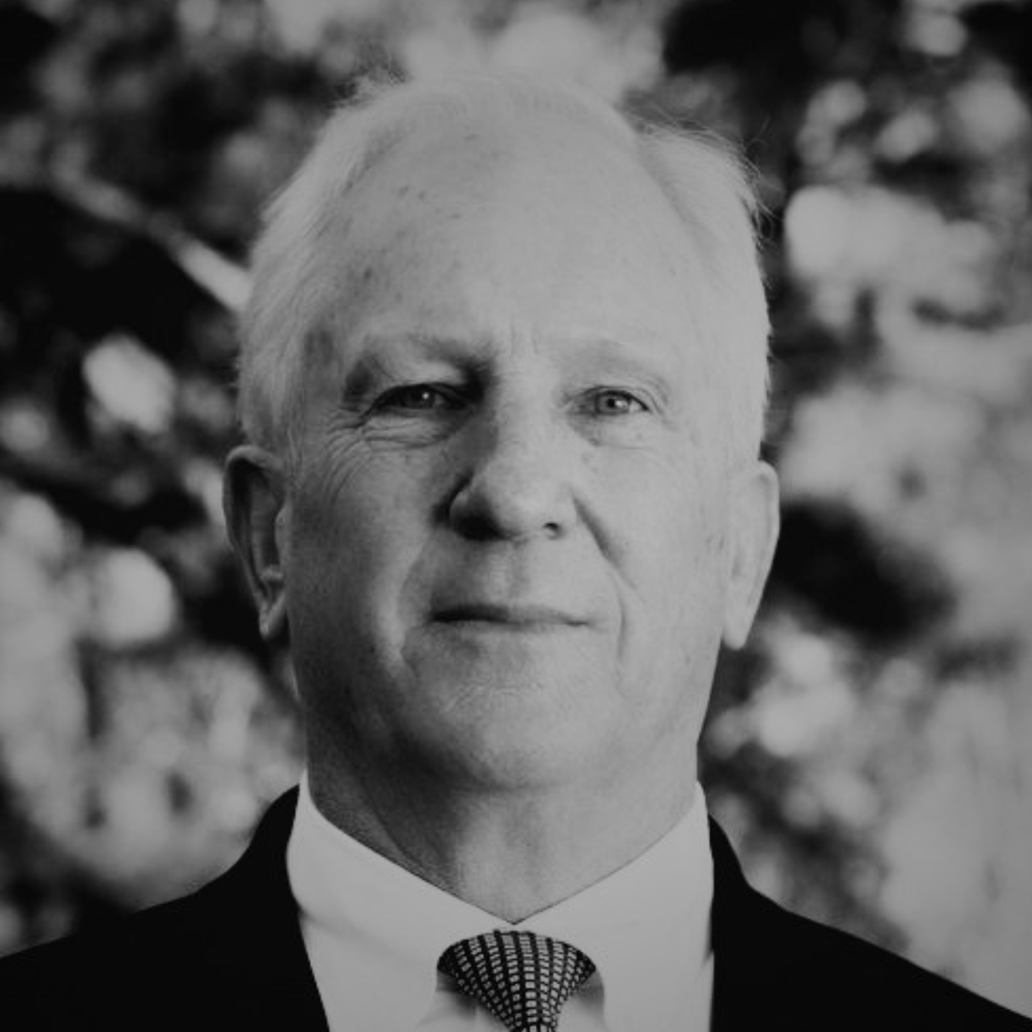 A senior man with white hair wearing a black suit, white shirt, and patterned tie, standing outdoors with trees in the background.