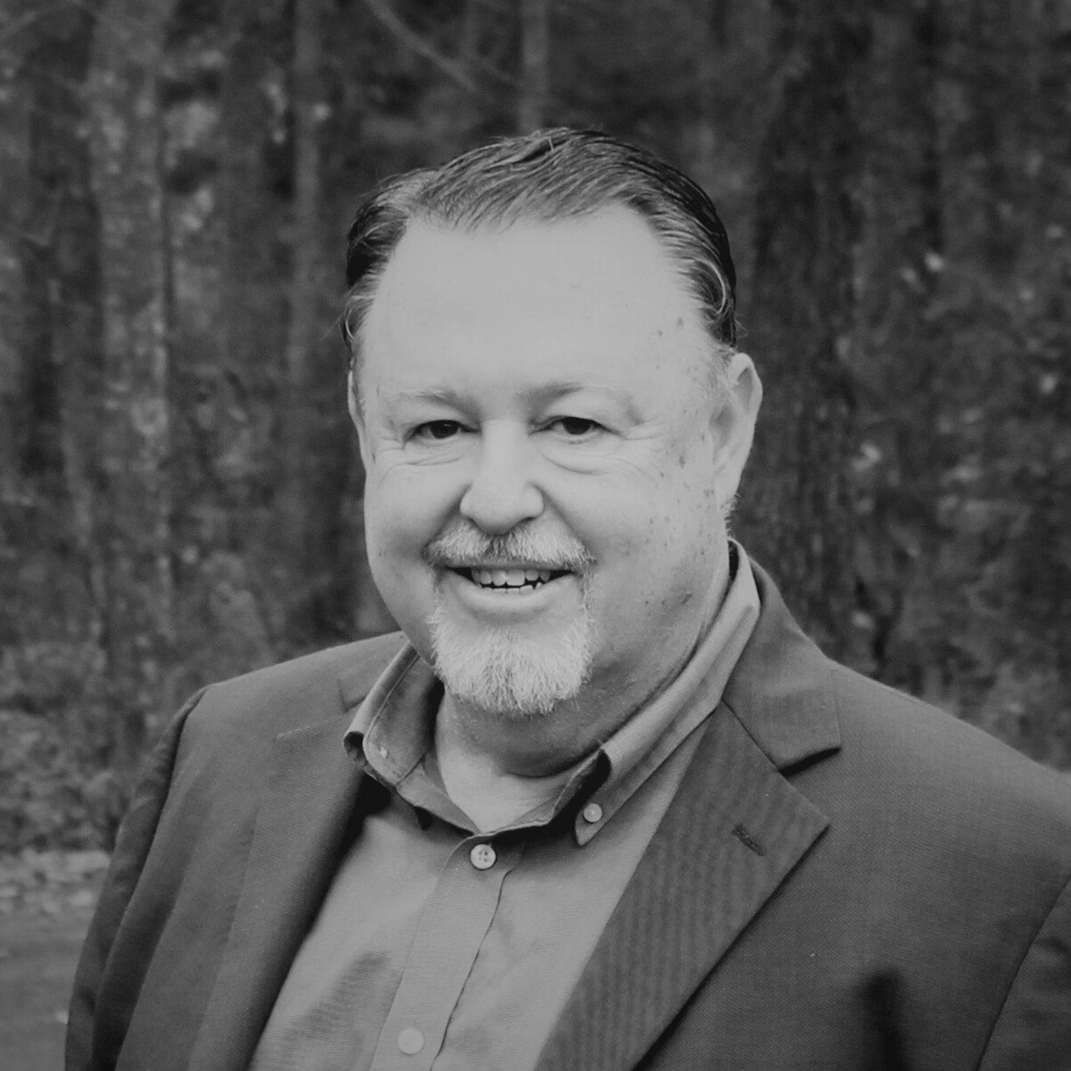 A smiling man with a beard and mustache wearing a light blue blazer over a blue and white checkered shirt, standing outdoors with trees in the background.
