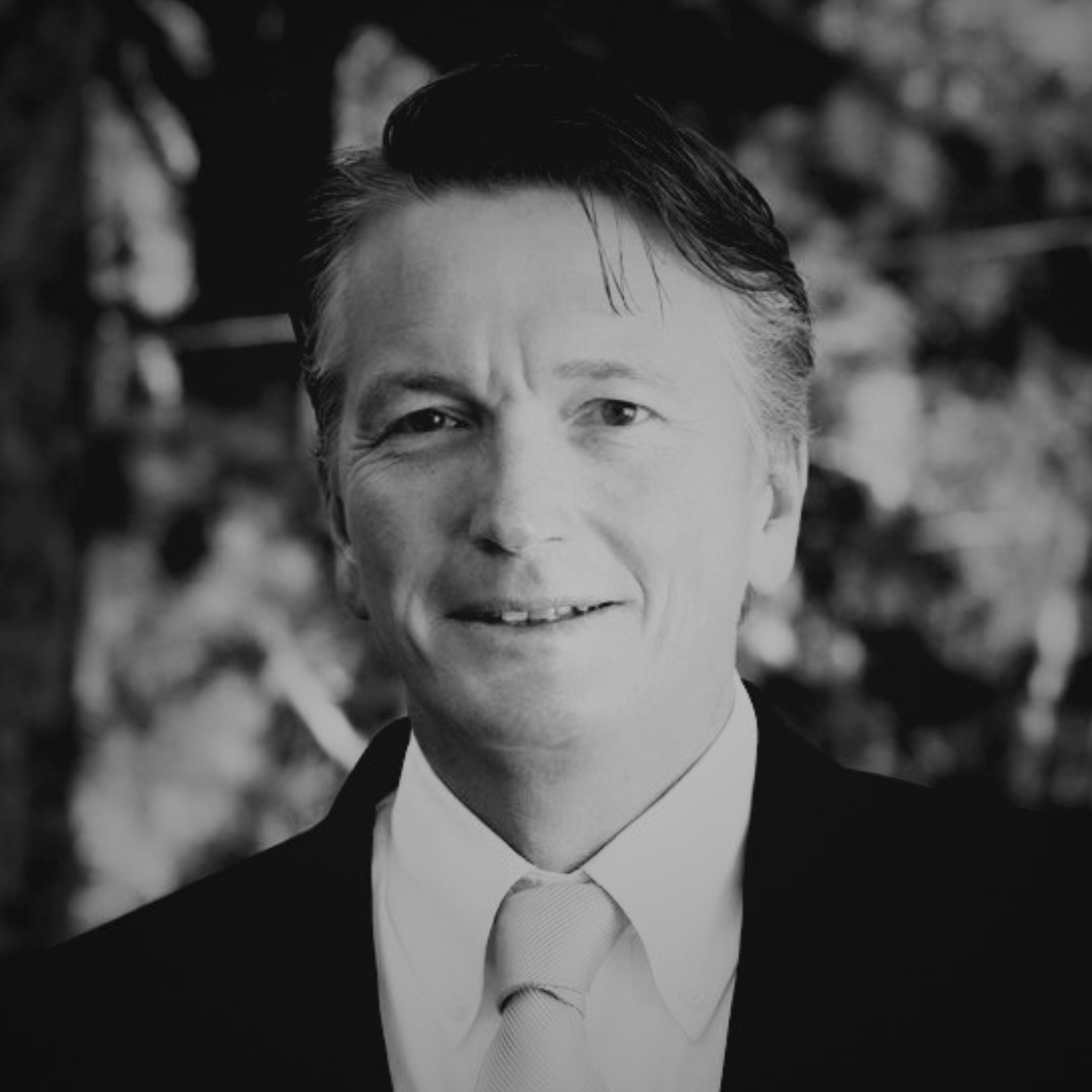 A man in a black suit with a light blue tie and white shirt smiling outdoors, with trees in the background.