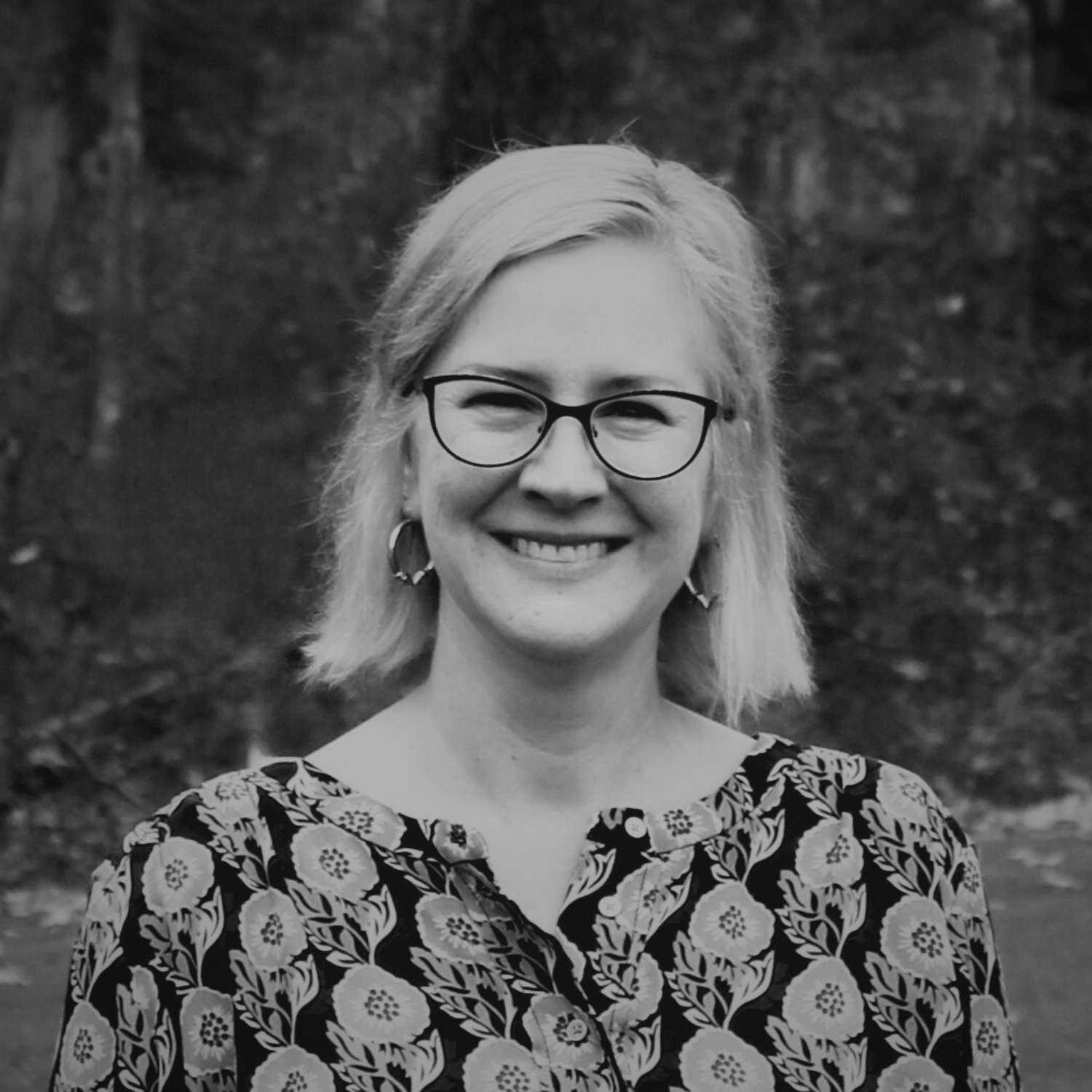 A woman with short, wavy blonde hair, glasses, and earrings, smiling outdoors with a blurred green background.