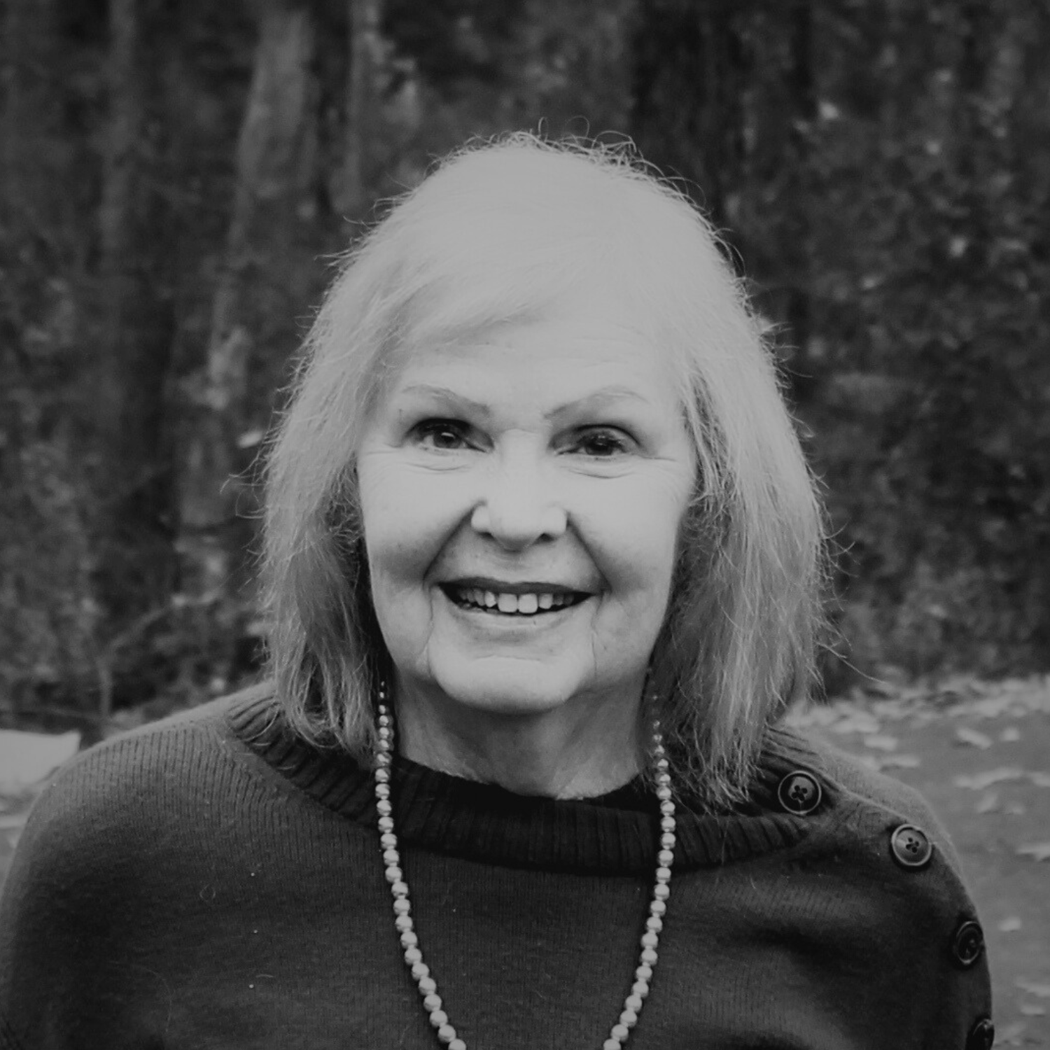 An elderly woman with gray hair, wearing a red sweater, gray pants, a gold necklace, and a white wristwatch, standing outdoors among trees with autumn foliage, smiling at the camera.