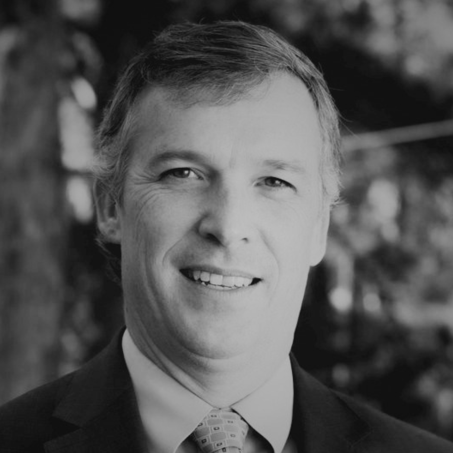 A middle-aged man wearing a dark suit jacket, light blue dress shirt, and a patterned yellow tie, standing outdoors with trees in the background.