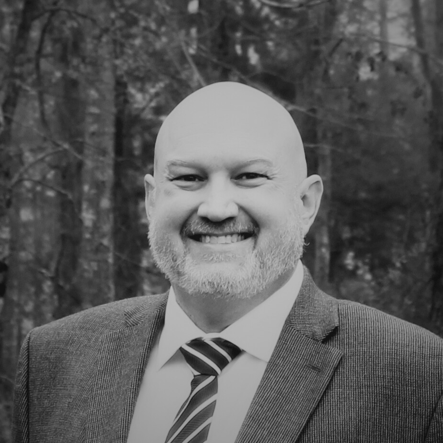 A man with a bald head and gray beard smiling outdoors, wearing a gray suit, white shirt, and striped tie, with trees in the background.