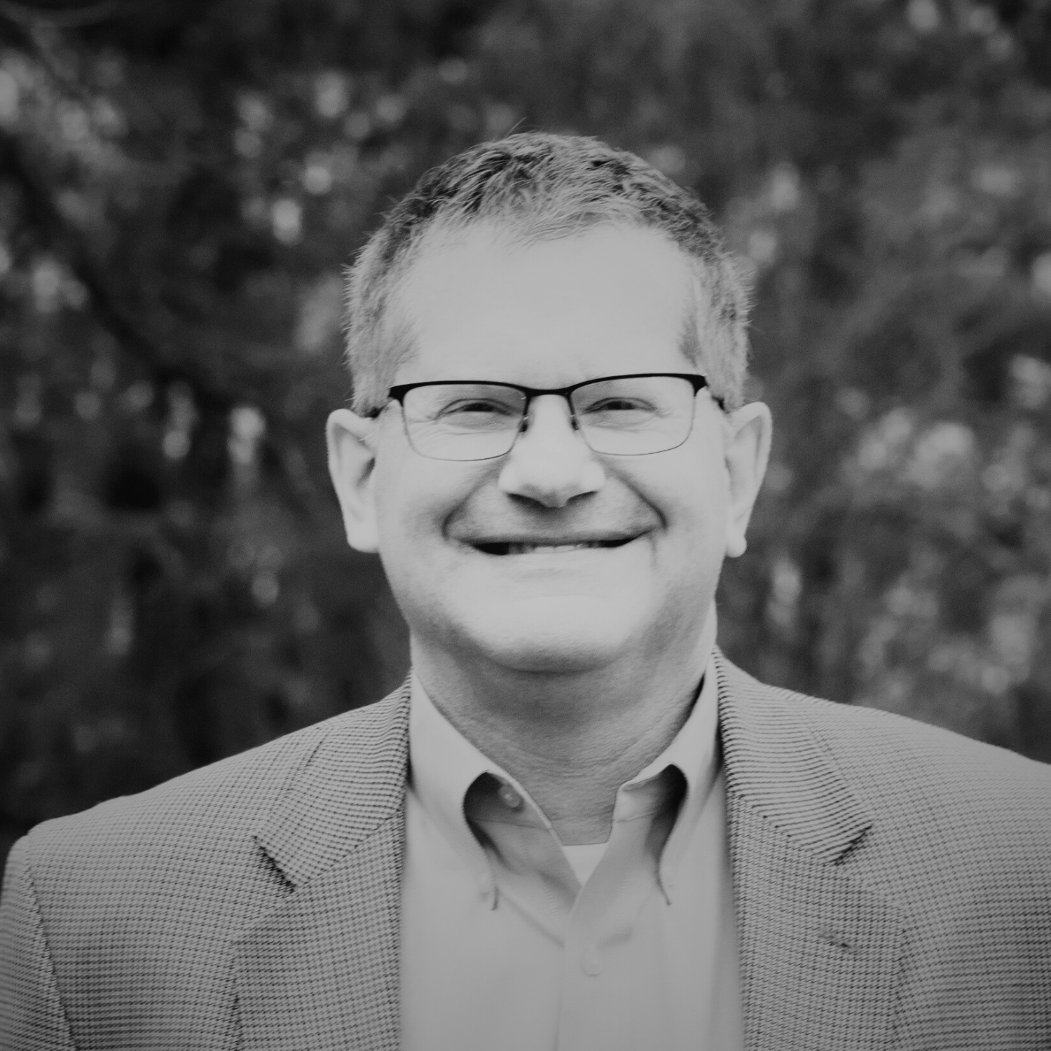 A smiling man in glasses wearing a gray plaid blazer and light blue shirt standing outdoors in front of trees.