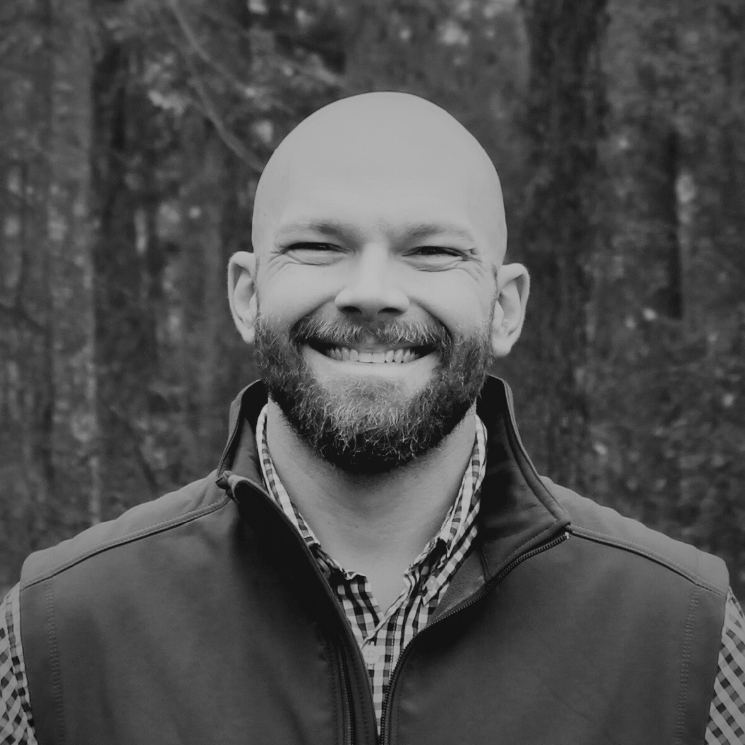 A smiling man with a beard and bald head, wearing a navy suit, white shirt, and a red-striped bow tie, standing outdoors with trees and autumn leaves in the background.