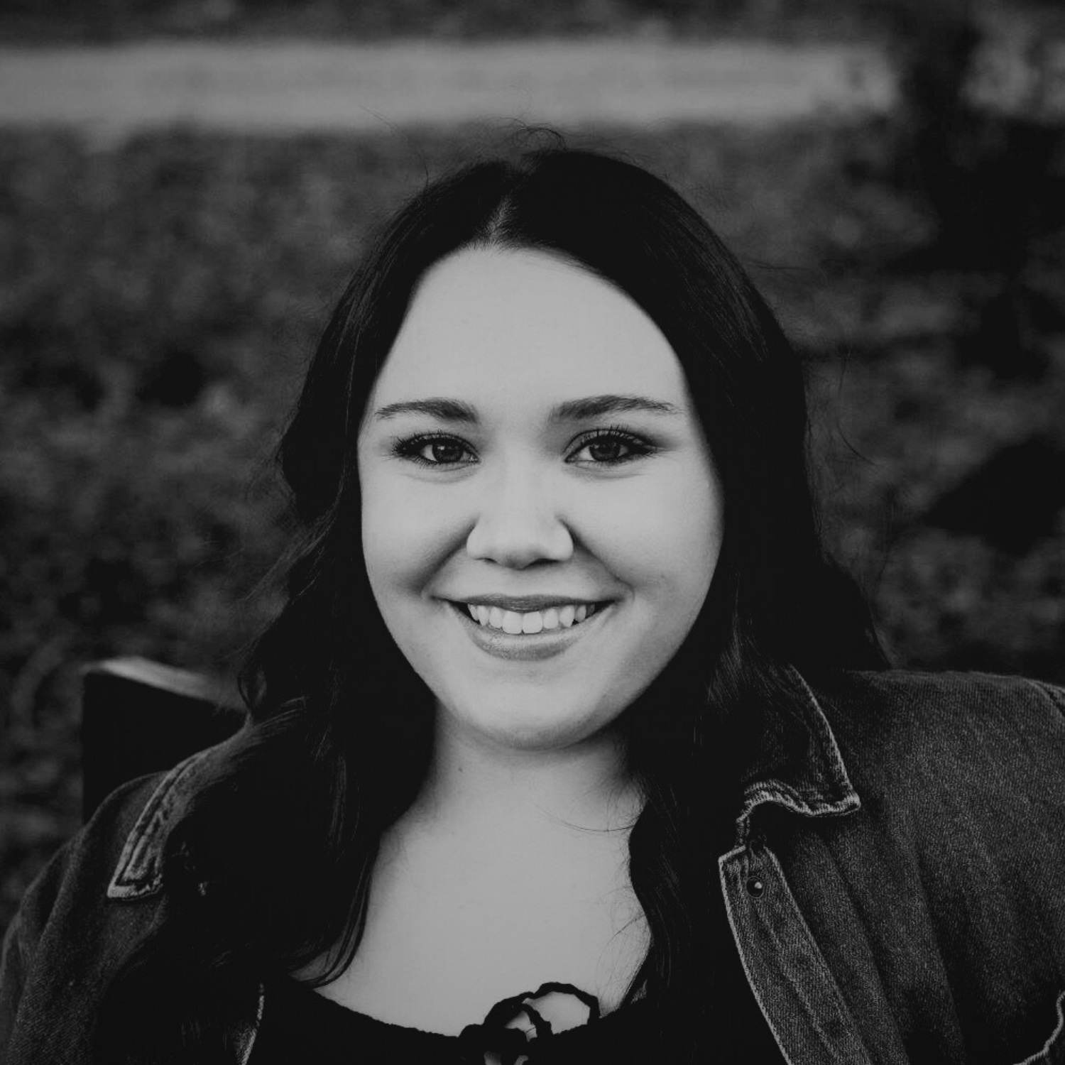 Smiling young woman with long brown hair, blue eyes, wearing a blue denim jacket and a black top, outdoors with blurred trees in the background.