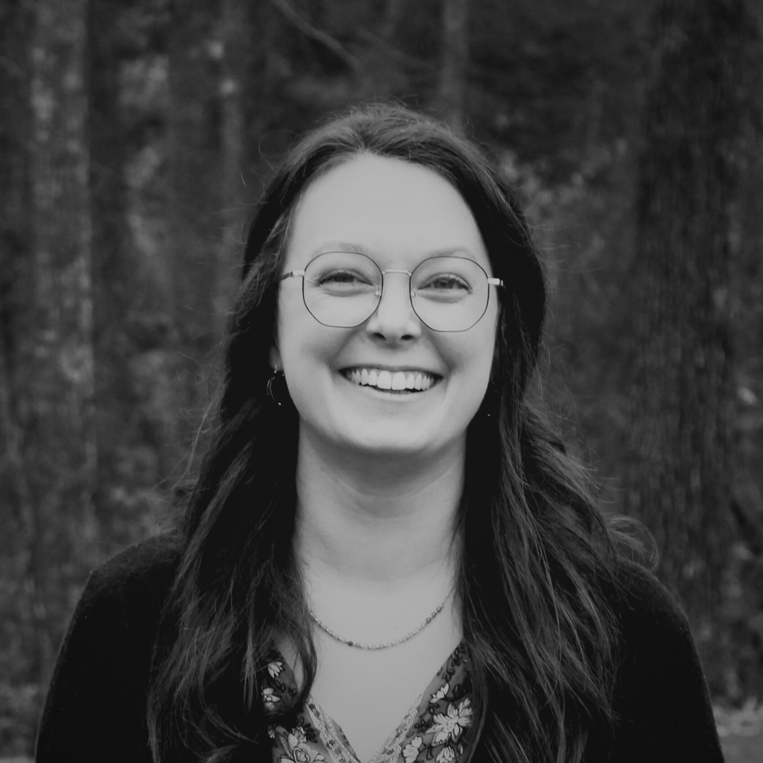 A woman with shoulder-length brown hair and bangs smiling outdoors in front of trees, wearing a navy floral dress and a light pink cardigan.