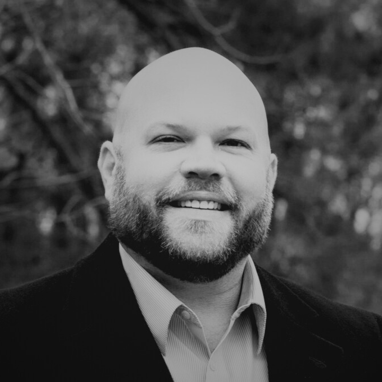 A bald man with a beard smiling outdoors in front of trees, wearing a black coat and a light-colored collared shirt.