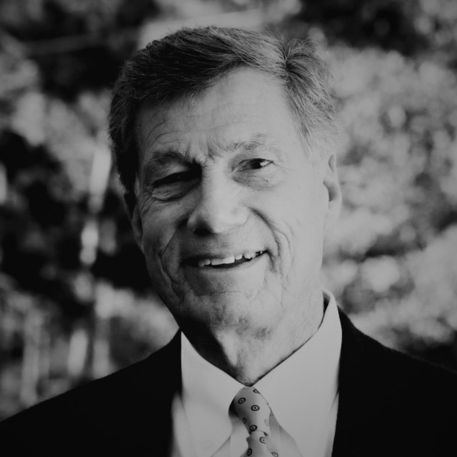 A smiling older man in a black suit, white shirt, and a beige polka dot tie, standing outdoors with trees and a clear sky in the background.