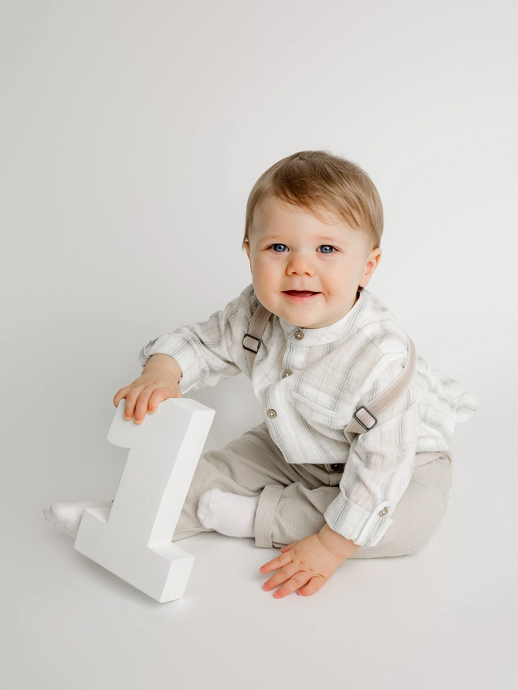 Pieni poika istuu lattialla, hymyilee kameraan, pitää kädessään puista isoa ykköstä.