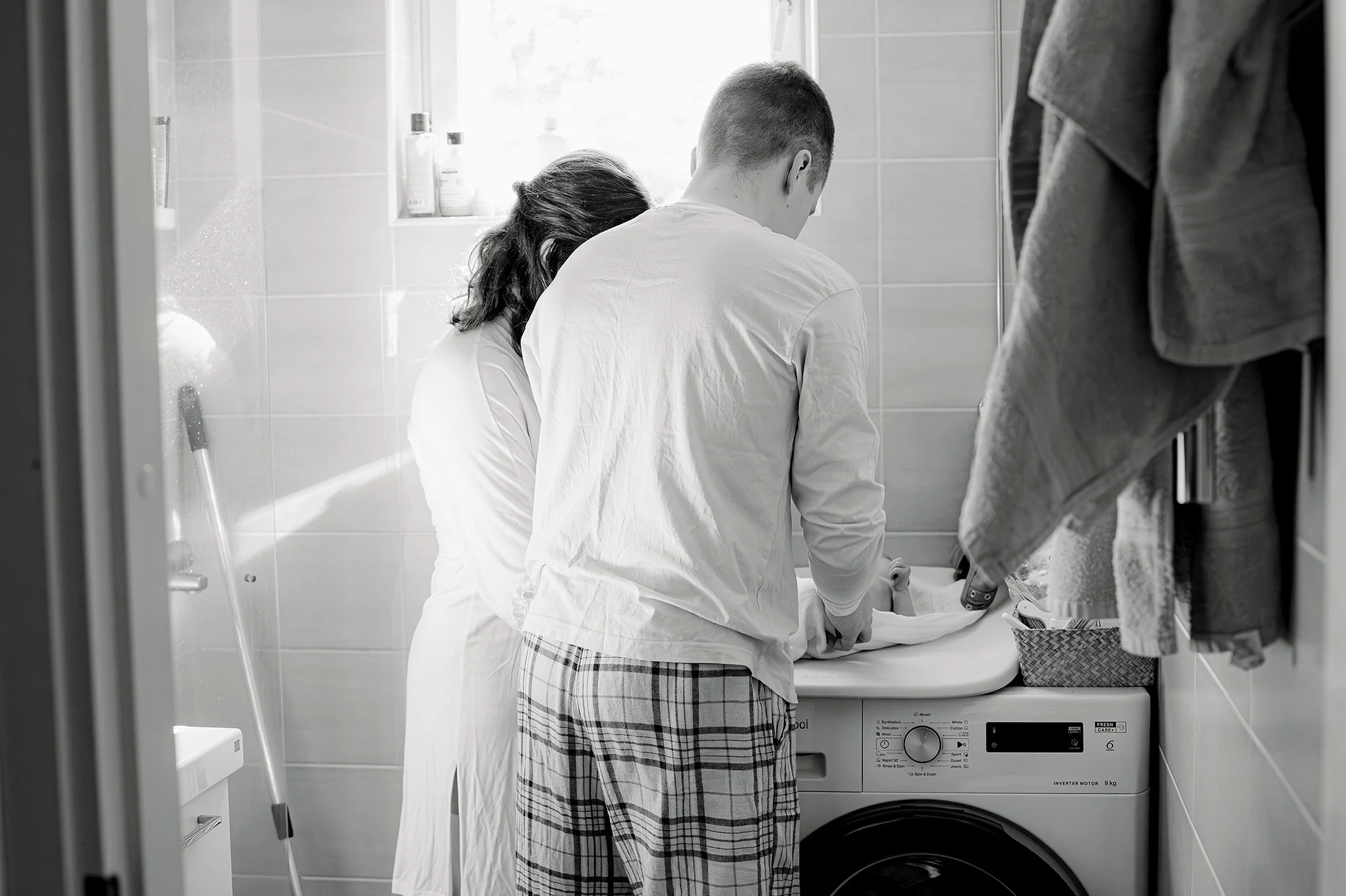 Äiti ja isä vaihtaa vaippaa vastasyntyneelle vauvalle kylpyhuoneessa, hoitopöytä on pesukoneen päällä. 